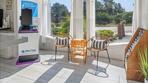 A check-in kiosk, two wicker chairs with cushions, a small table, and a view of greenery through large windows.