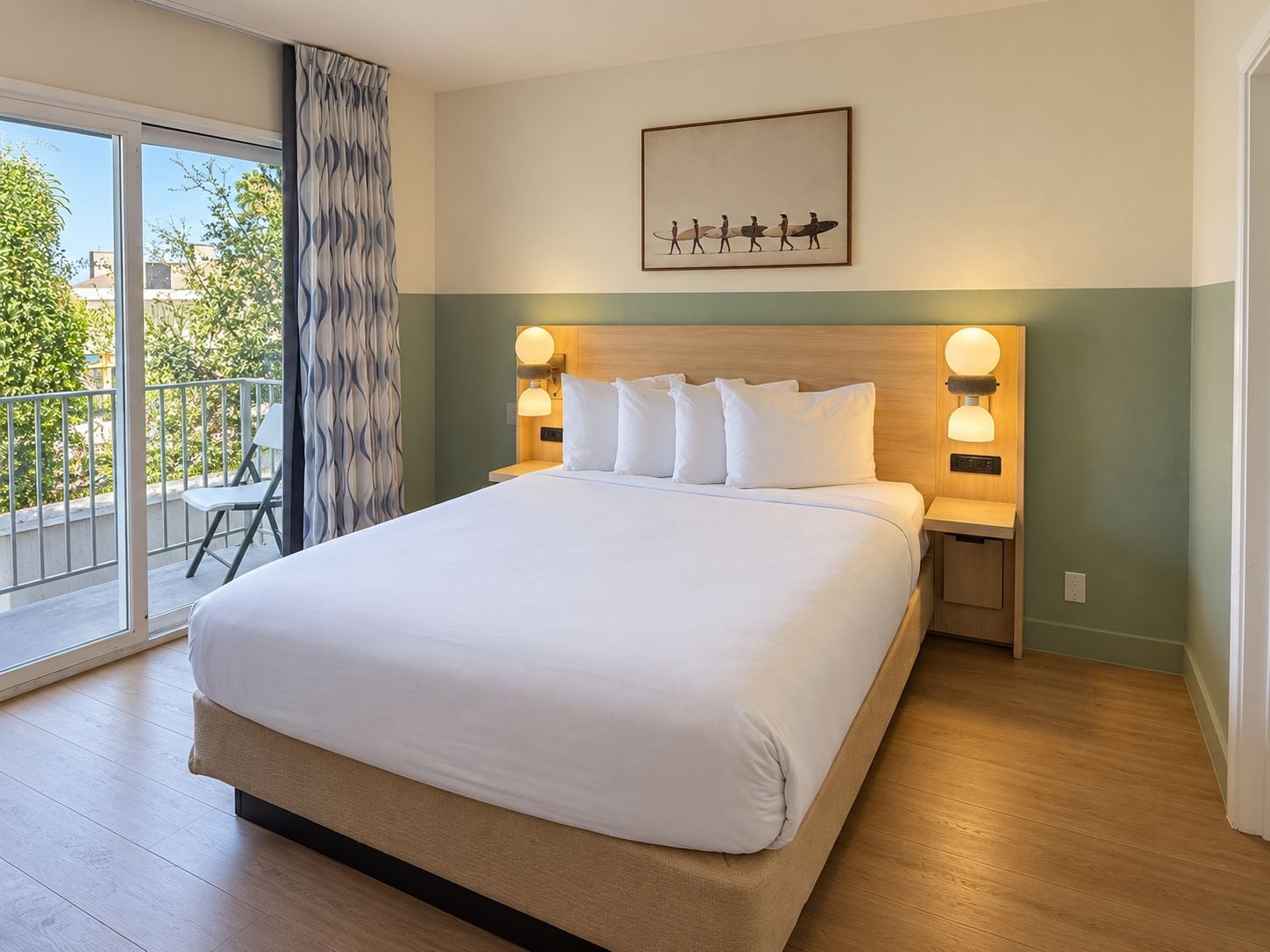 Habitación de hotel con una cama grande, dos mesitas de noche con lámparas, vistas al exterior y un cuadro en la pared.