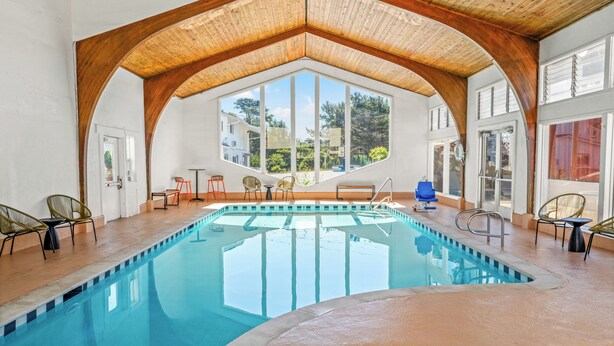 An indoor swimming pool with a wooden ceiling, clear glass walls, and multiple seating areas.