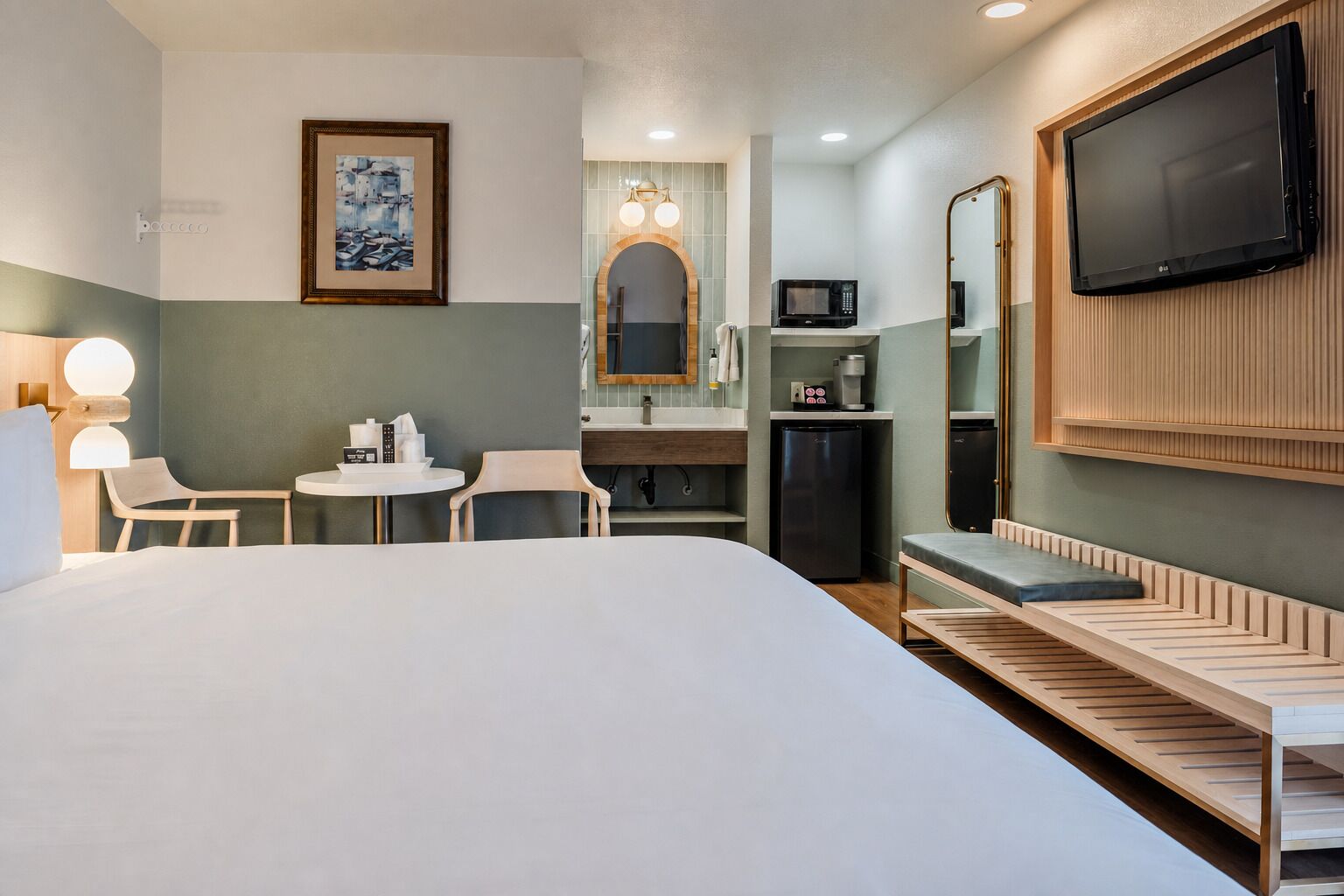 A hotel room with a large bed, a desk with a chair, a small round table, a microwave, a refrigerator, a TV mounted on the wall, and a framed picture on the wall.