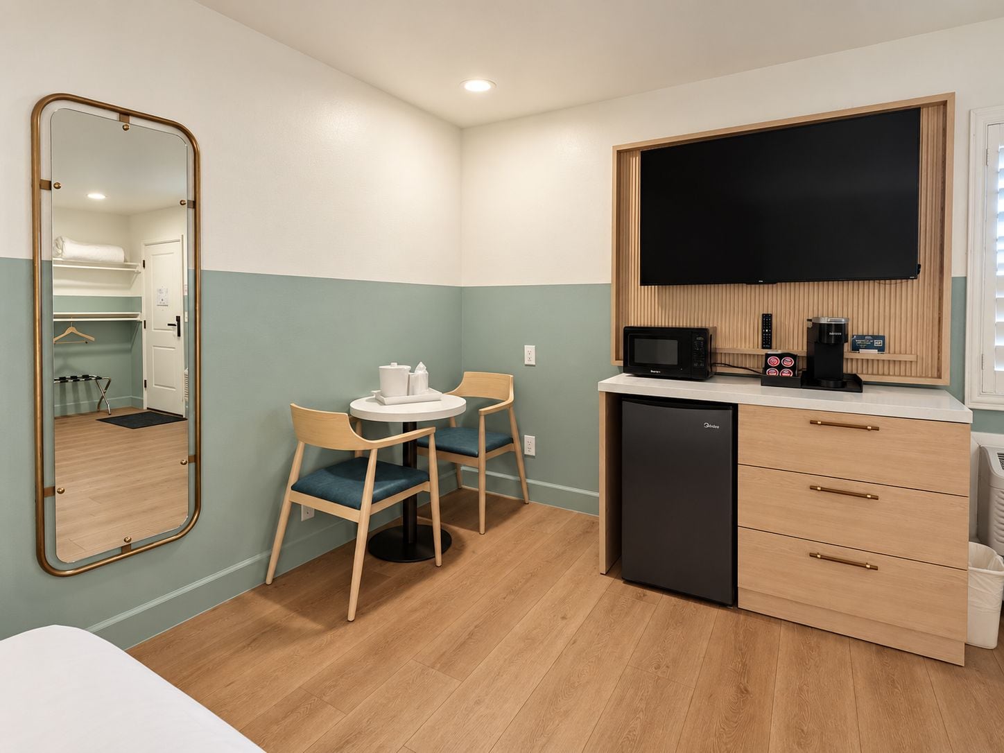 A compact kitchenette with a microwave, coffee maker, and a small refrigerator. There is a round table with two chairs and a mirror on the wall.