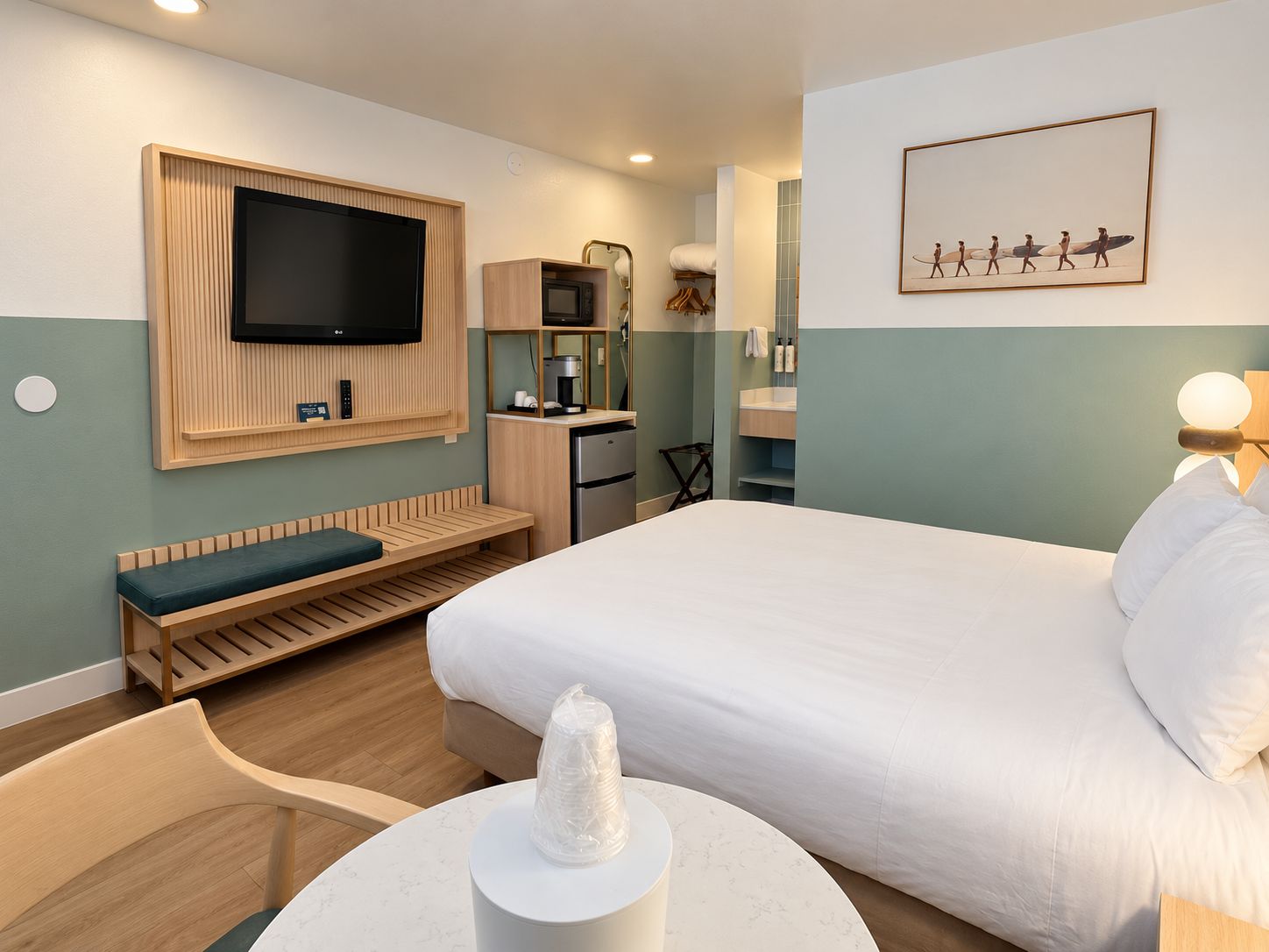 A hotel room with a bed, a TV mounted on the wall, a desk with a chair, a small kitchenette, and a bathroom.