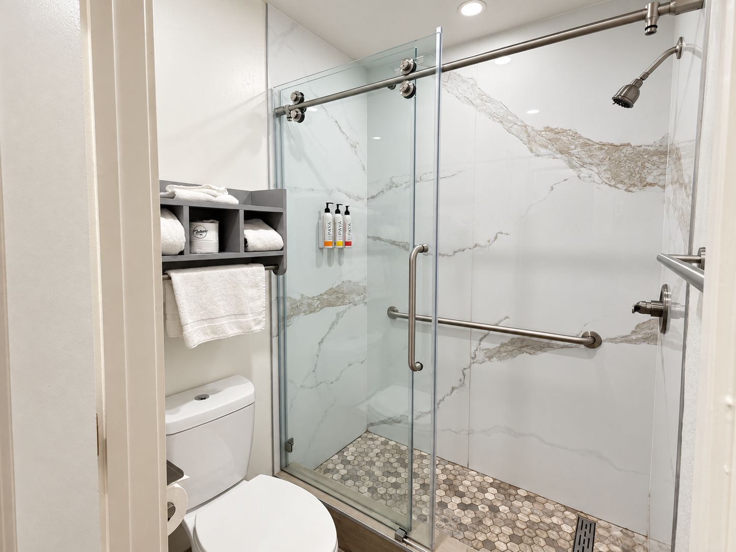 A modern bathroom with a glass-enclosed shower, marble walls, a toilet, and a shelf with toiletries.