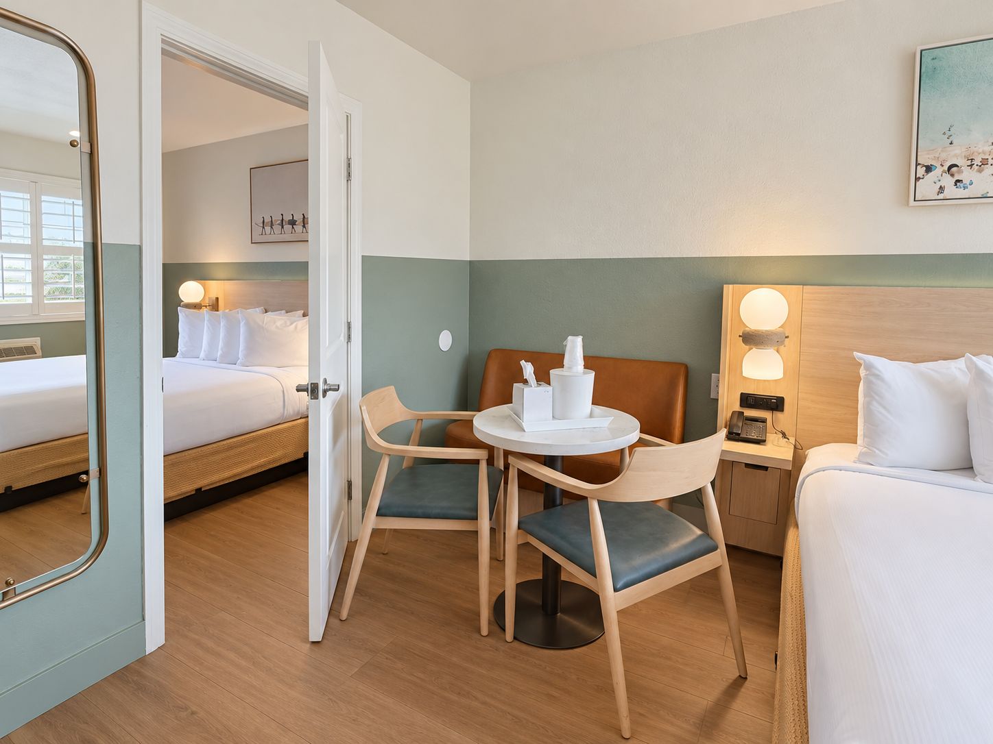 A hotel room with a bed, a table with chairs, a nightstand, and a wall-mounted lamp.