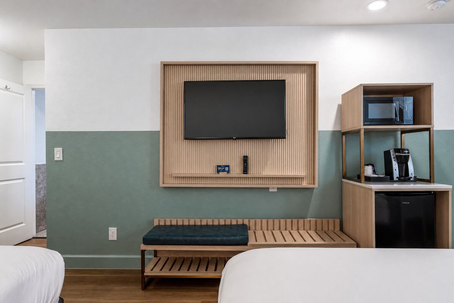 A hotel room with a flat-screen TV mounted on the wall, a wooden bench, a coffee machine, and a mini-fridge.