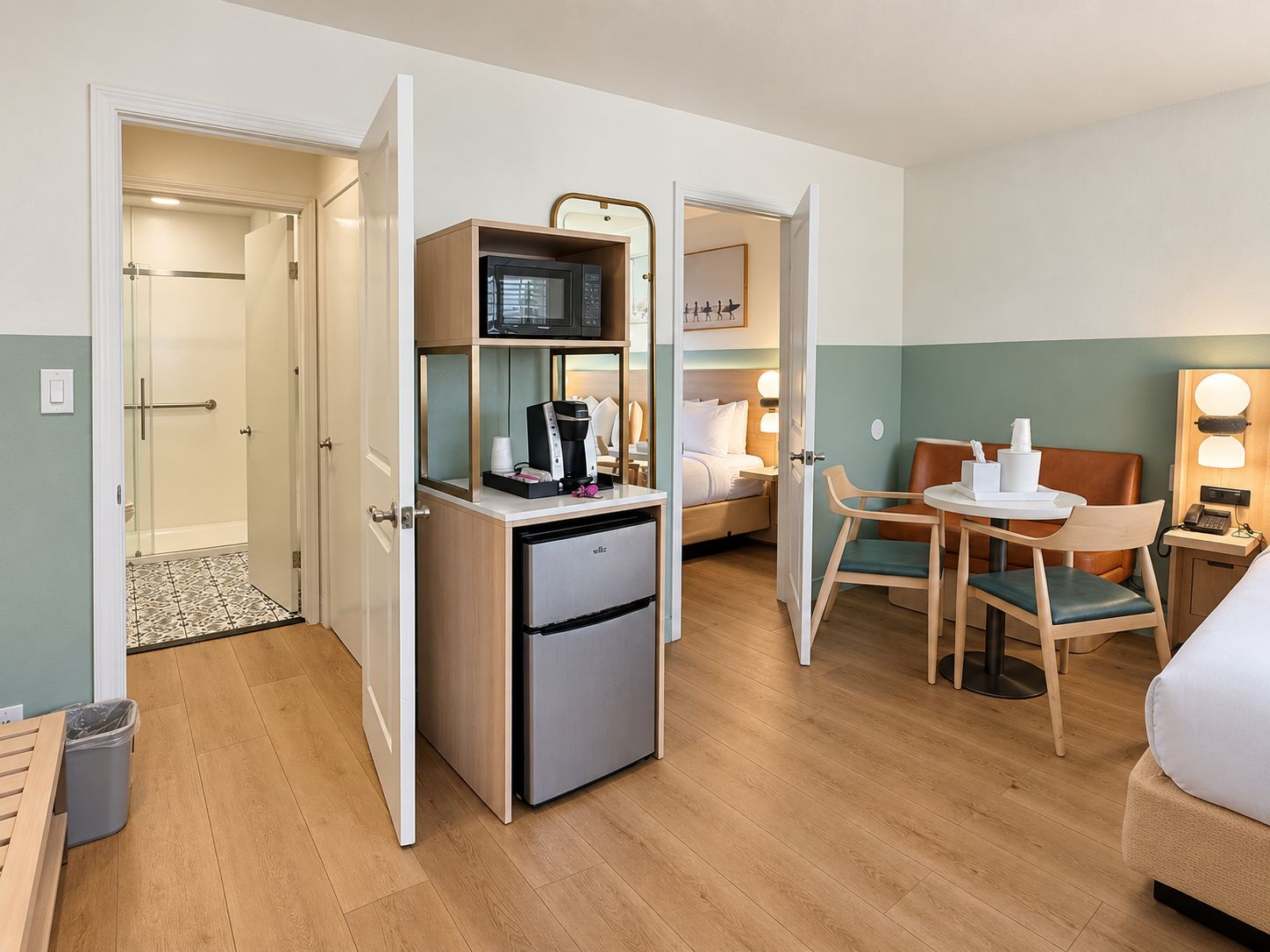 A compact hotel room with a kitchenette, a bed, a seating area, and a shower.