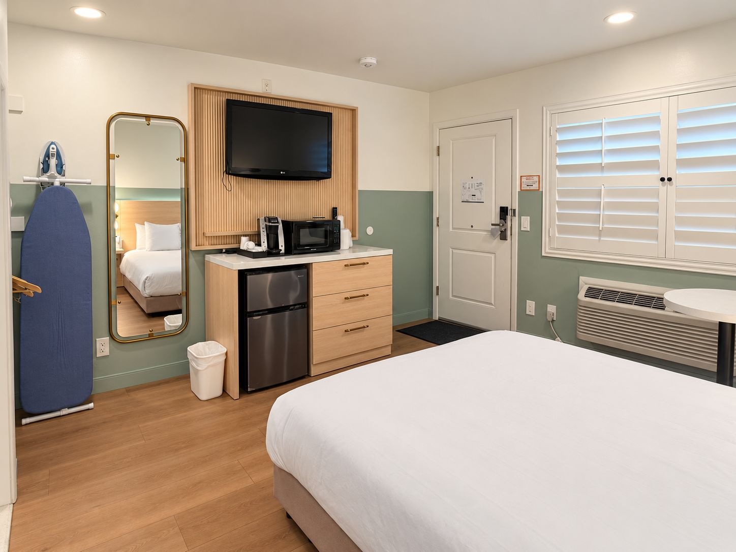 Habitación de hotel con una cama, un televisor montado en un panel de madera, un microondas, un refrigerador pequeño, un espejo, una puerta con ventana y un equipo de aire acondicionado de pared.