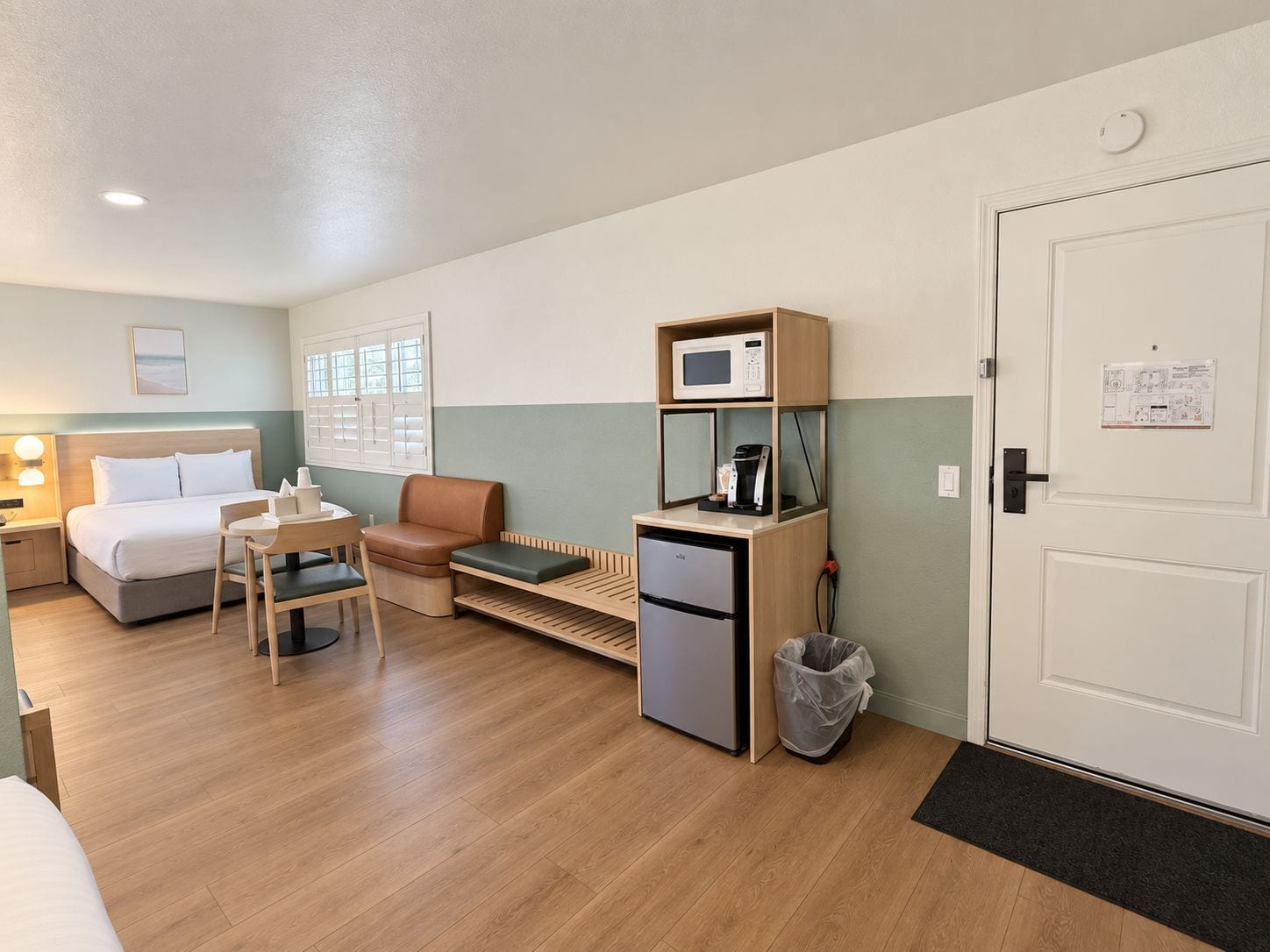 A compact hotel room with a bed, a seating area with a table and chairs, a kitchenette with a microwave and coffee machine, and a door with a notice.