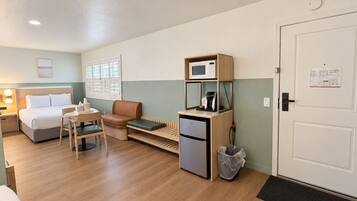 A compact hotel room with a bed, a seating area with a table and chairs, a kitchenette with a microwave and coffee machine, and a door with a notice.