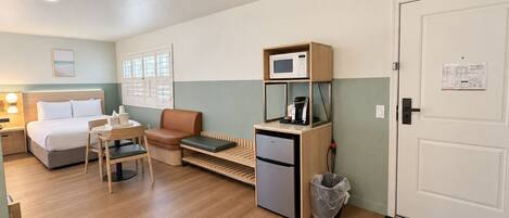 A compact hotel room with a bed, a seating area with a table and chairs, a kitchenette with a microwave and coffee machine, and a door with a notice.