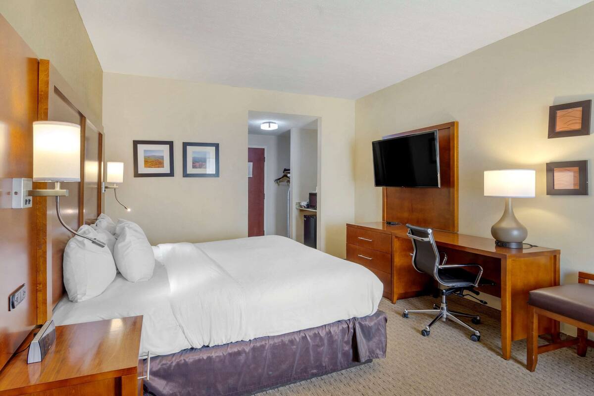 A hotel room with a bed, desk, chair, TV, and a small bathroom visible through an open door.