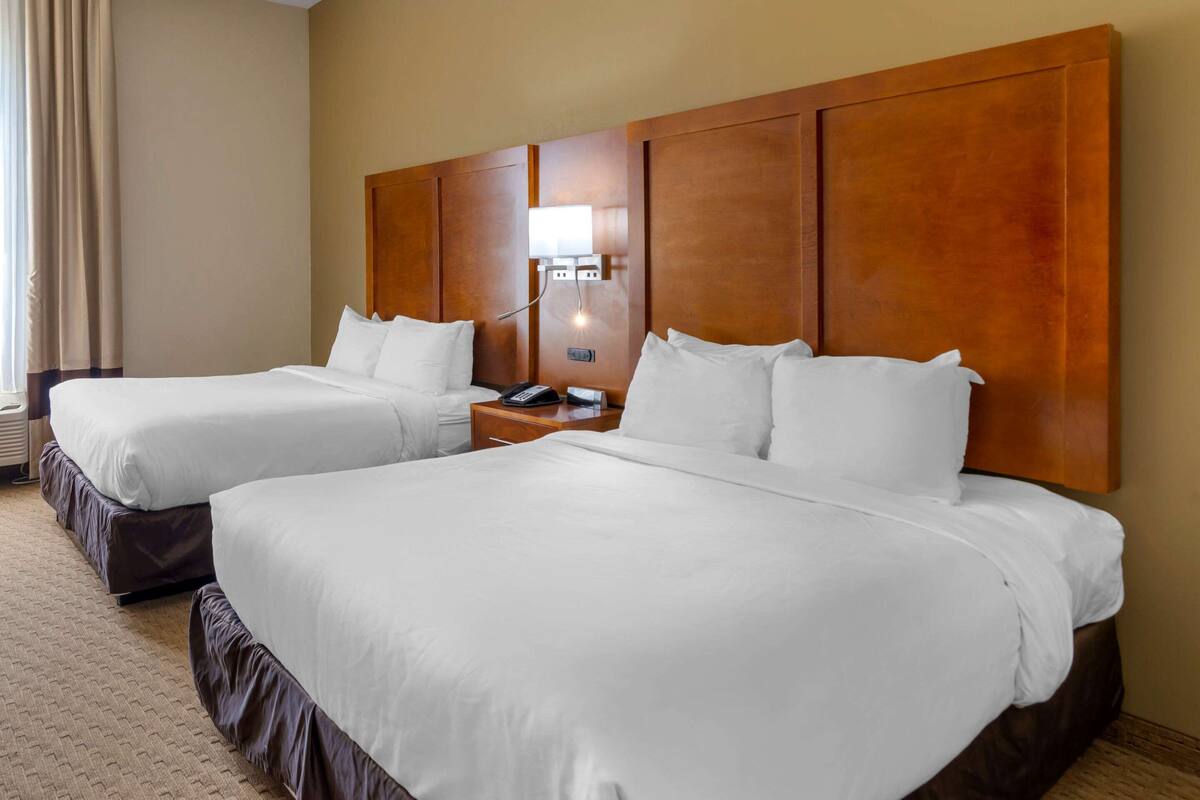 A hotel room with two beds, a wooden headboard, a bedside table with a lamp, and a window with curtains.