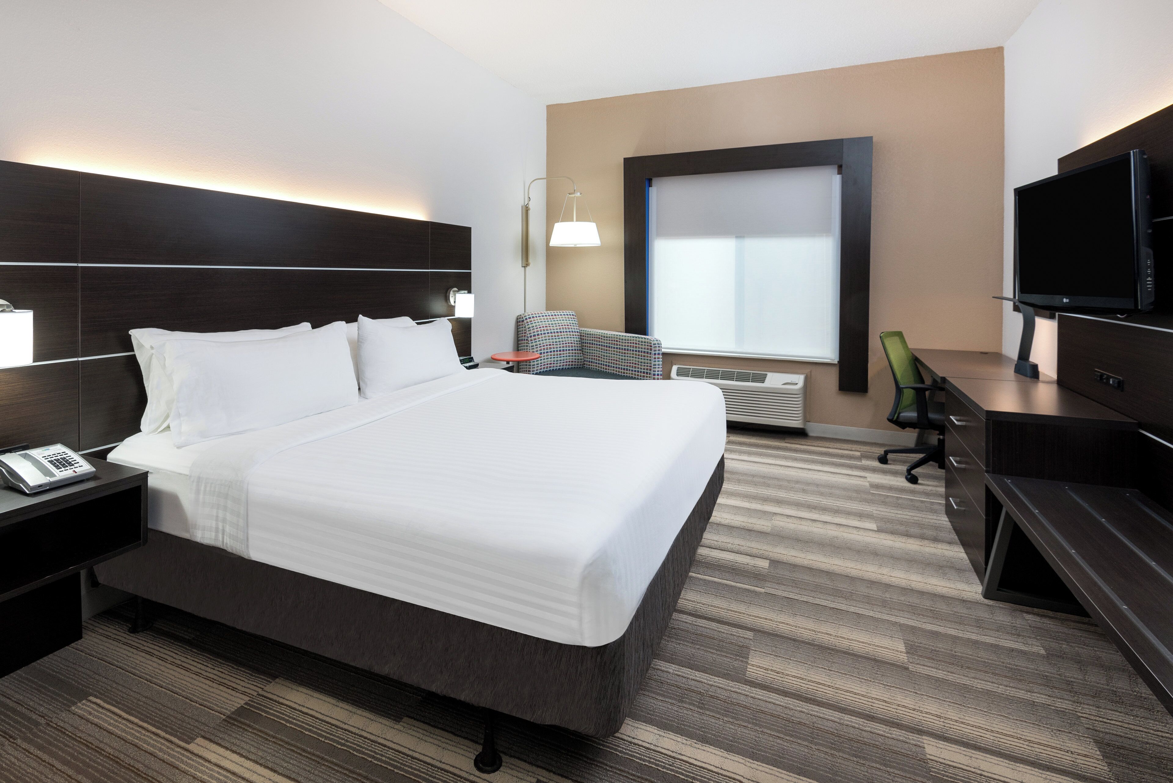 Habitación de hotel con una cama grande, un escritorio con computadora, televisor y ventana con persianas.