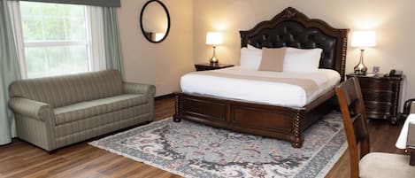 A hotel room with a large bed, a sofa, a chair, a nightstand with a lamp, and a window with curtains.
