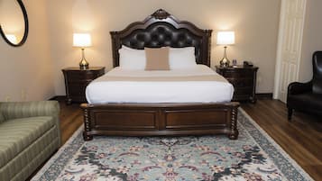 Una cama bien hecha con un cabecero oscuro, flanqueada por dos mesitas de noche con lámparas, una alfombra estampada y un sillón verde.