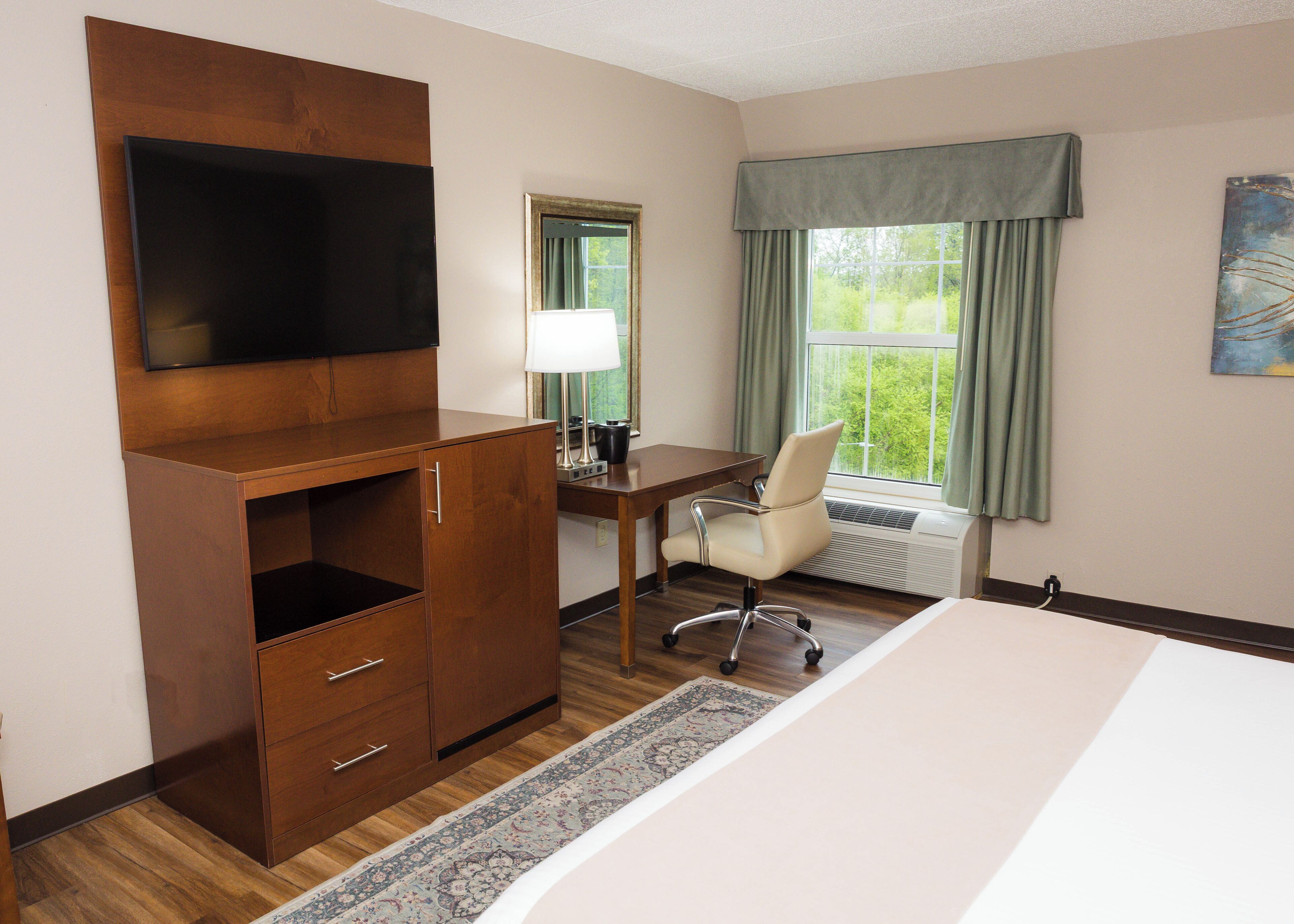 Una habitación de hotel con una cama, un escritorio con silla, un televisor montado en un mueble de madera, una ventana con cortinas y un radiador.