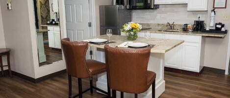 A modern kitchen with white cabinetry, a stainless steel refrigerator, a microwave, and a granite countertop. There are two brown leather bar stools and a vase with yellow flowers on the island.