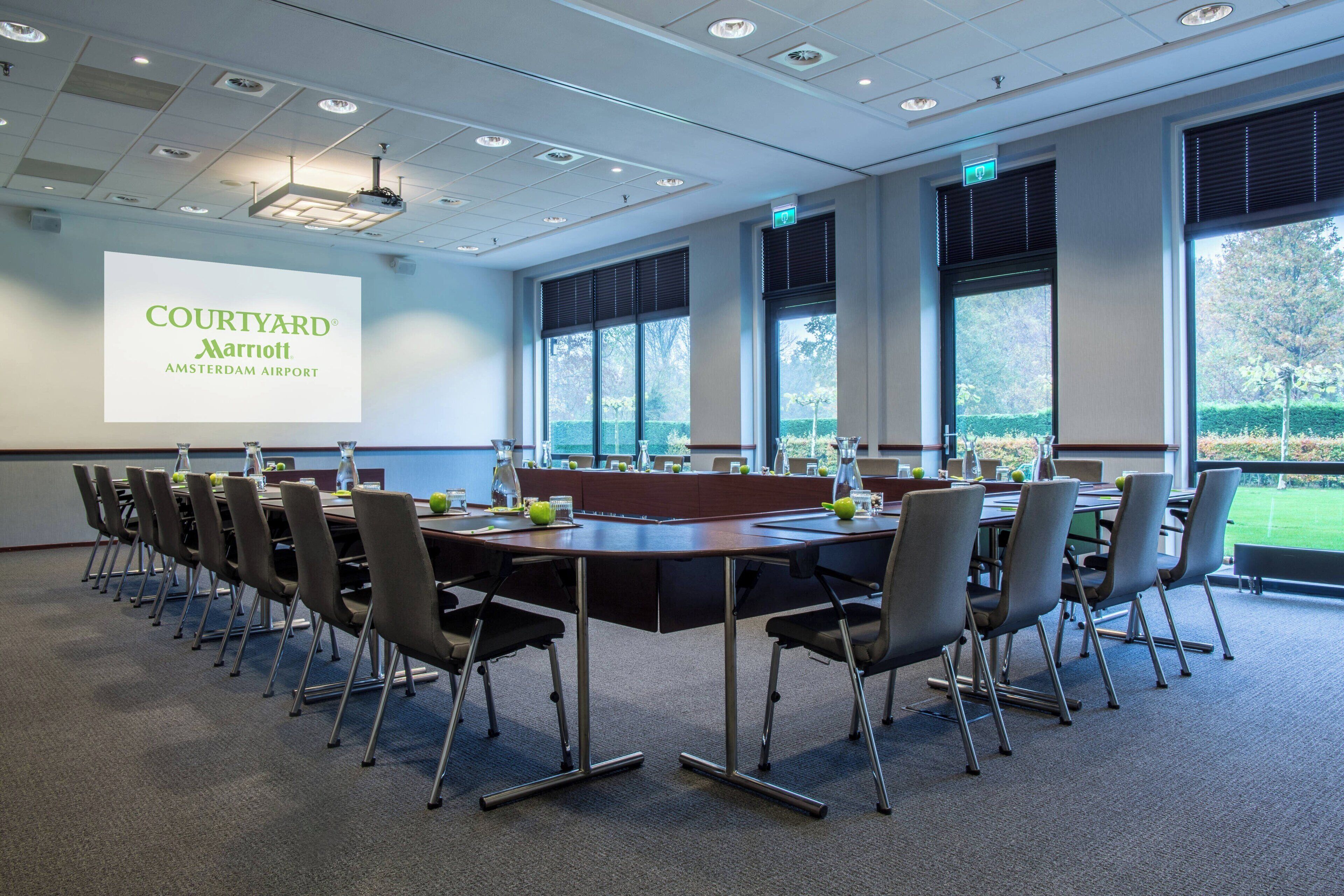 Een vergaderruimte met een lange tafel, stoelen, een projectiescherm waarop 'Courtyard Marriott Amsterdam Airport' staat, en grote ramen met uitzicht op het omliggende landschap.