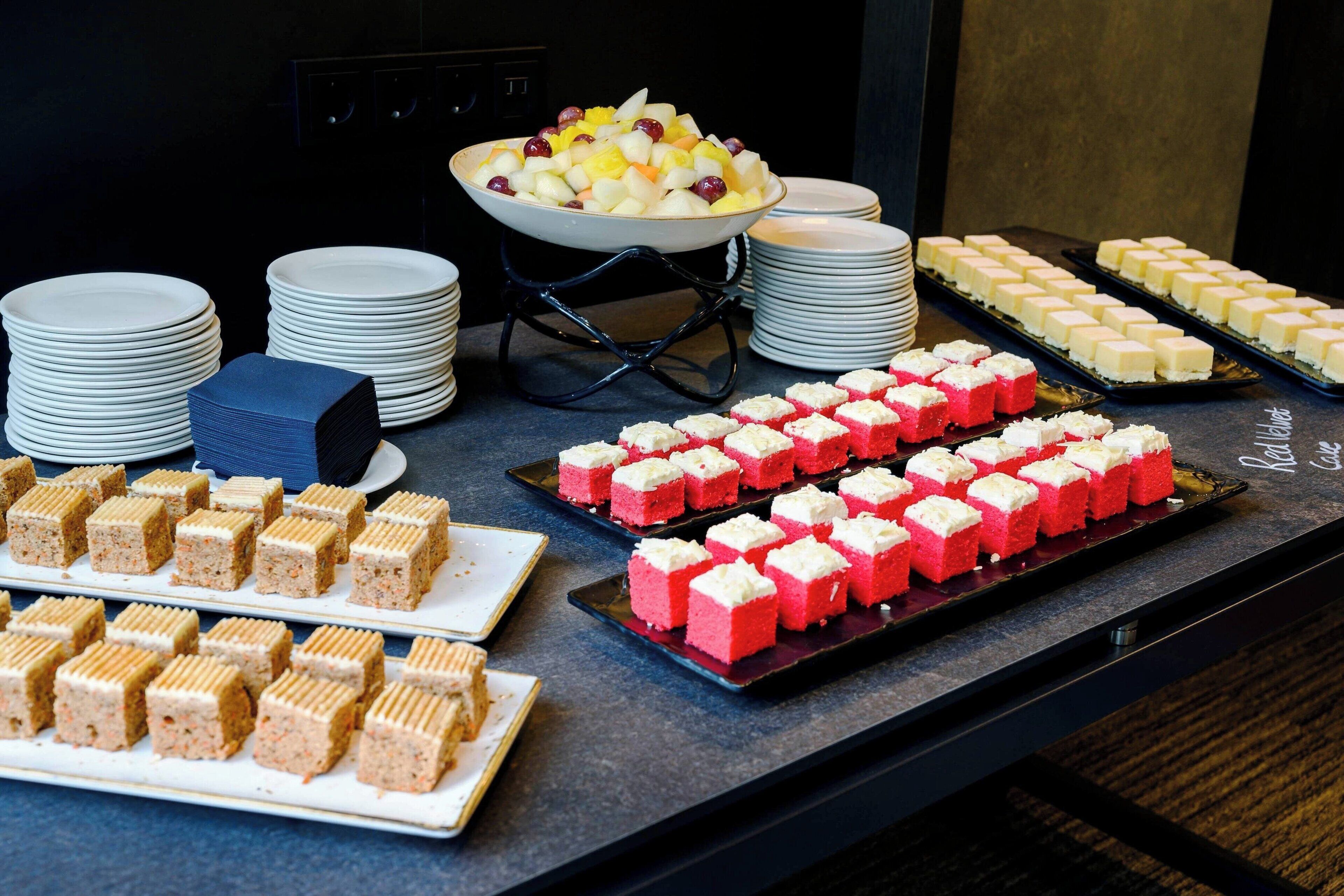 A buffet table with various food items including a bowl of fruit salad, trays of red and white desserts, and stacks of white plates.