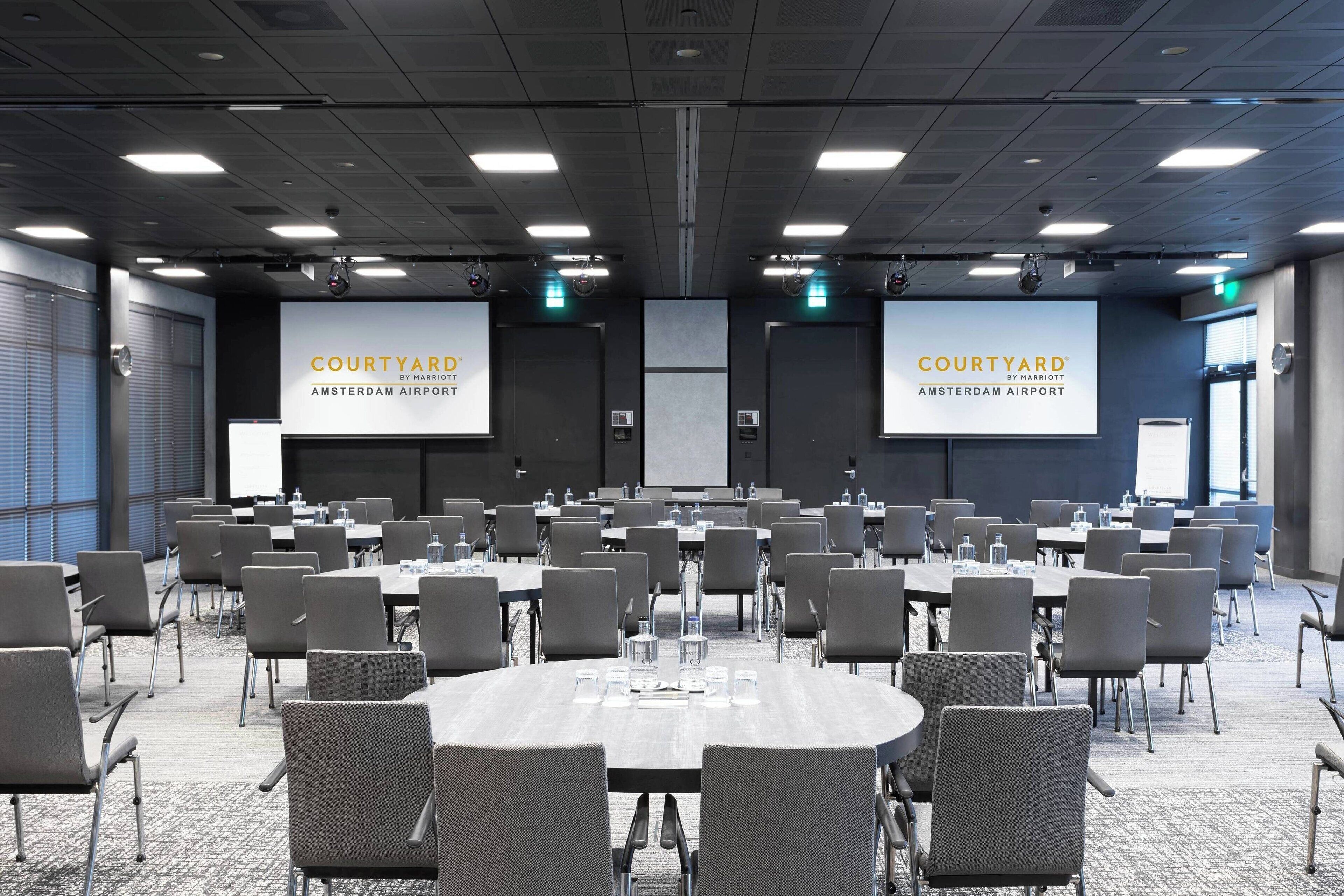 Ein Konferenzraum mit mehreren Tischen und Stühlen, großen Bildschirmen, auf denen 'Courtyard by Marriott Amsterdam Airport' zu sehen ist, und einem modernen, gut beleuchteten Innenraum.