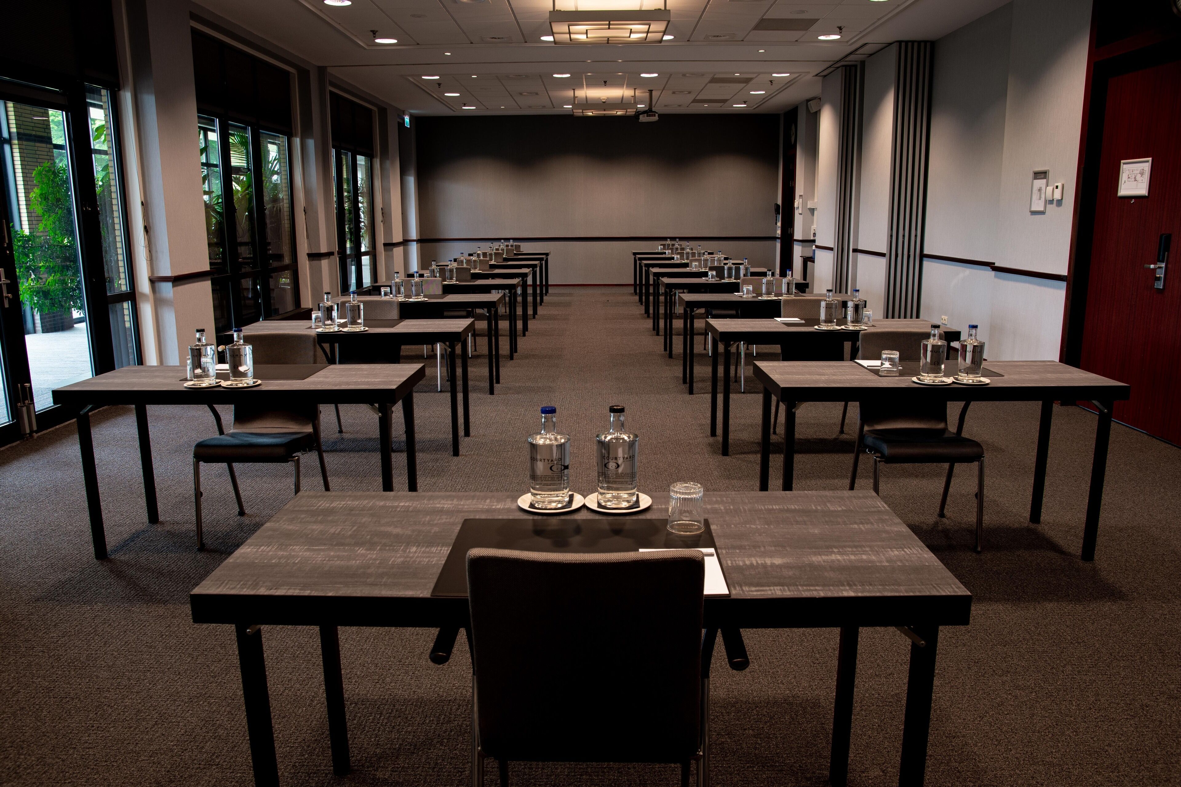 A conference room with multiple tables and chairs, equipped with water bottles and glasses, large windows, and a projector screen.