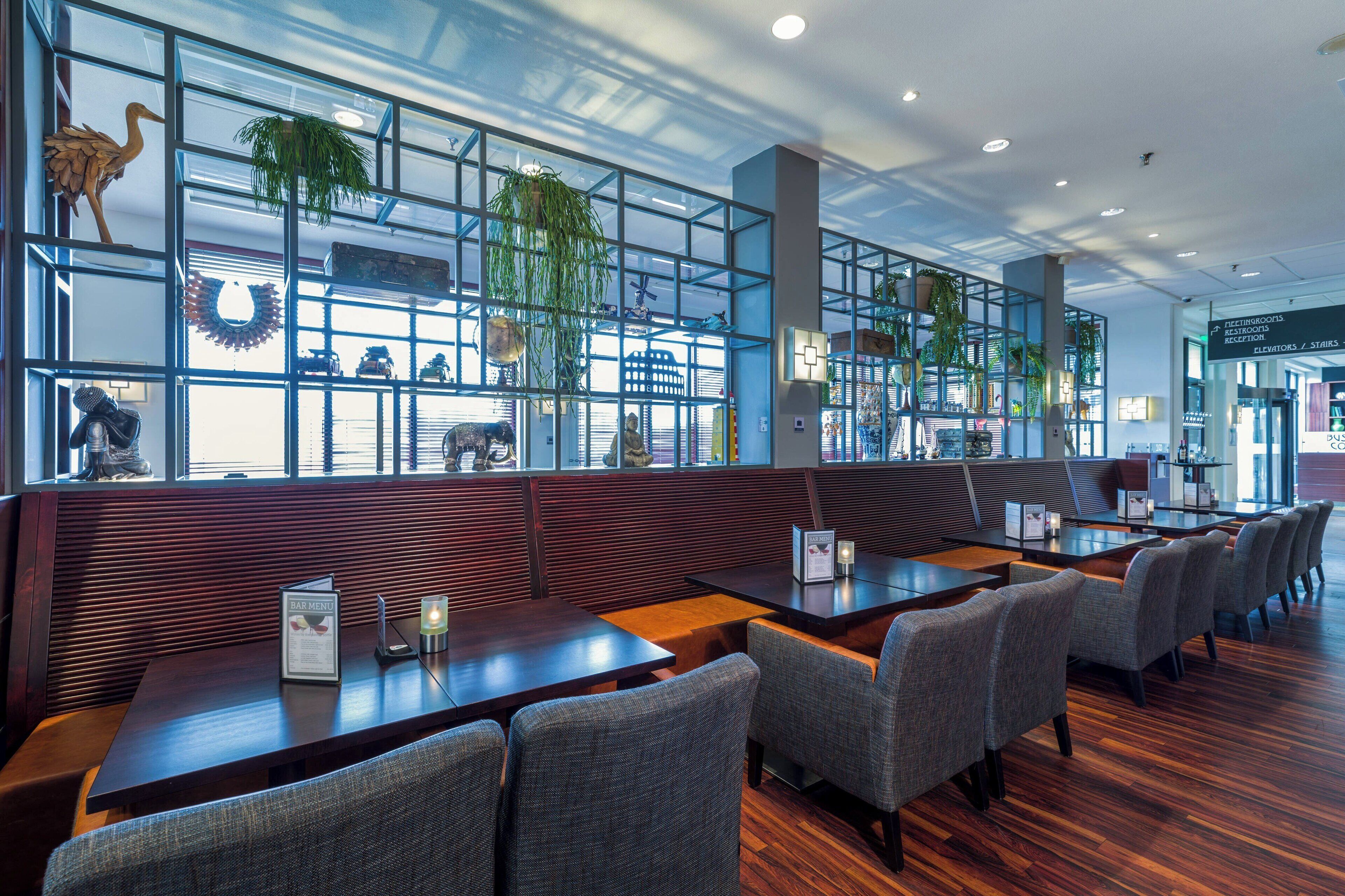 Ein modernes Restaurant mit Holzecken und Tischen, großen Fenstern mit Pflanzen und einer gut sichtbaren Speisekarte.