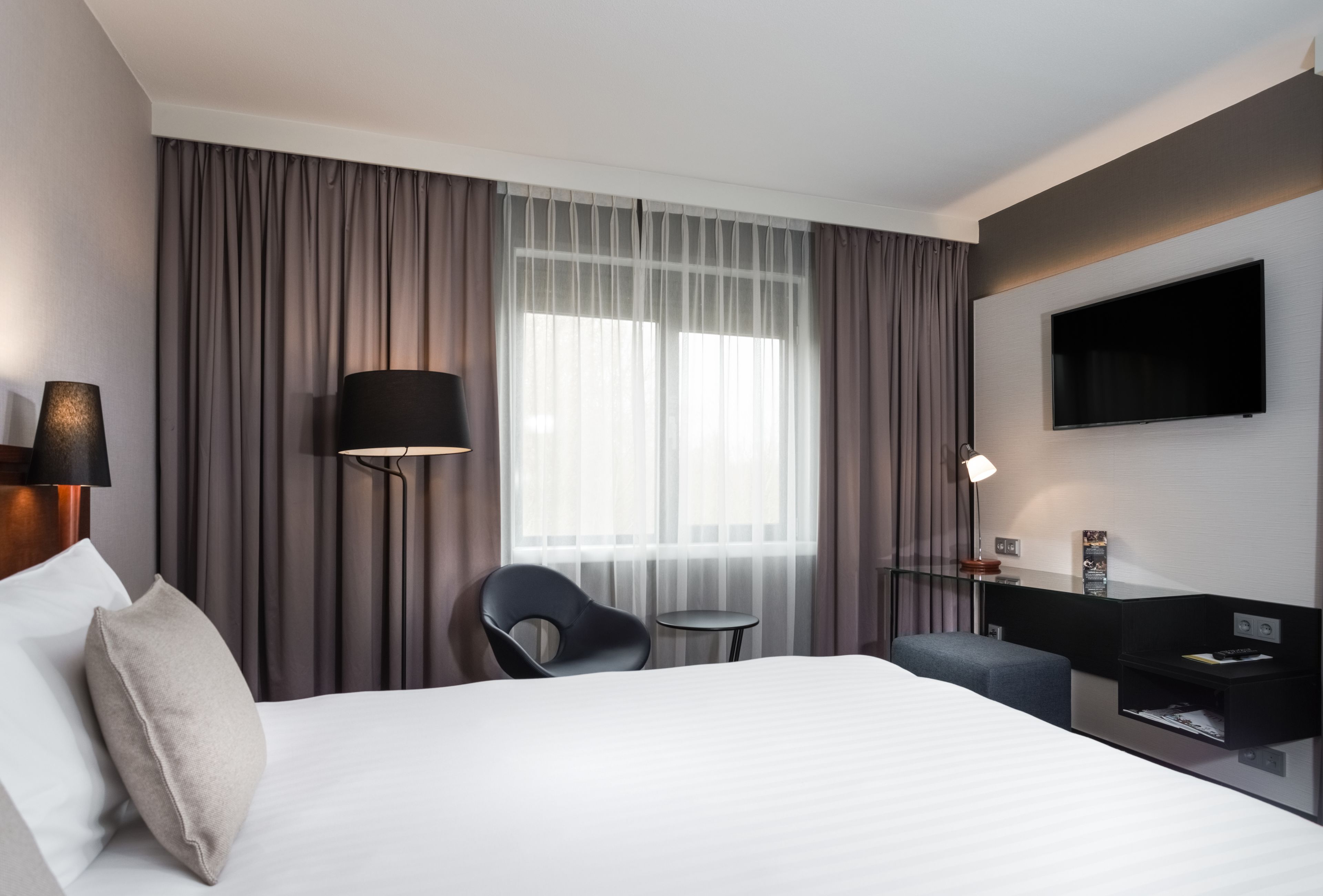 Una habitación de hotel moderna con una cama grande, un escritorio, una silla, una mesita, un televisor y una ventana con cortinas.