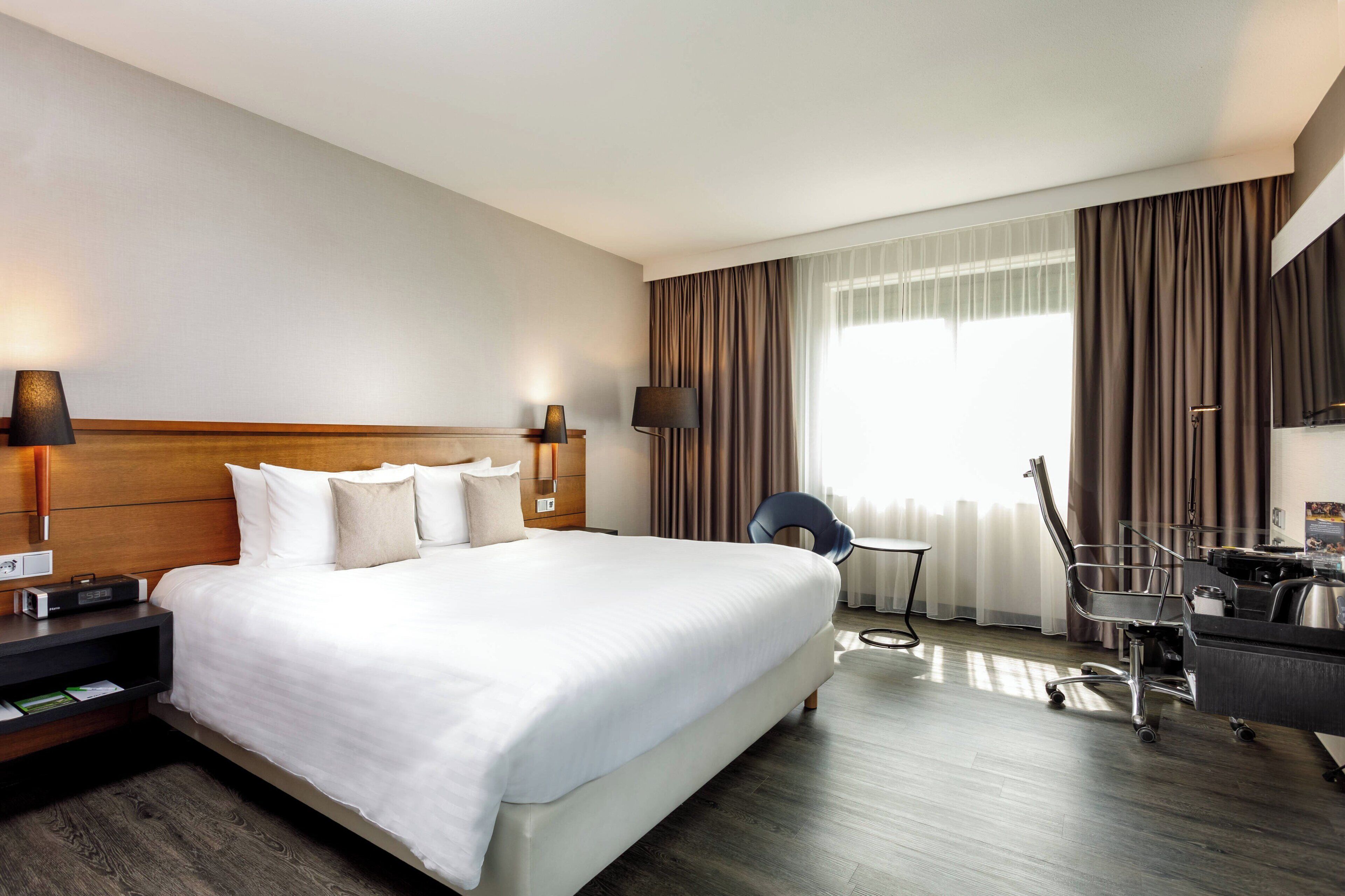Une chambre d’hôtel avec un grand lit, des tables de chevet, un bureau avec une chaise et une fenêtre avec des rideaux.