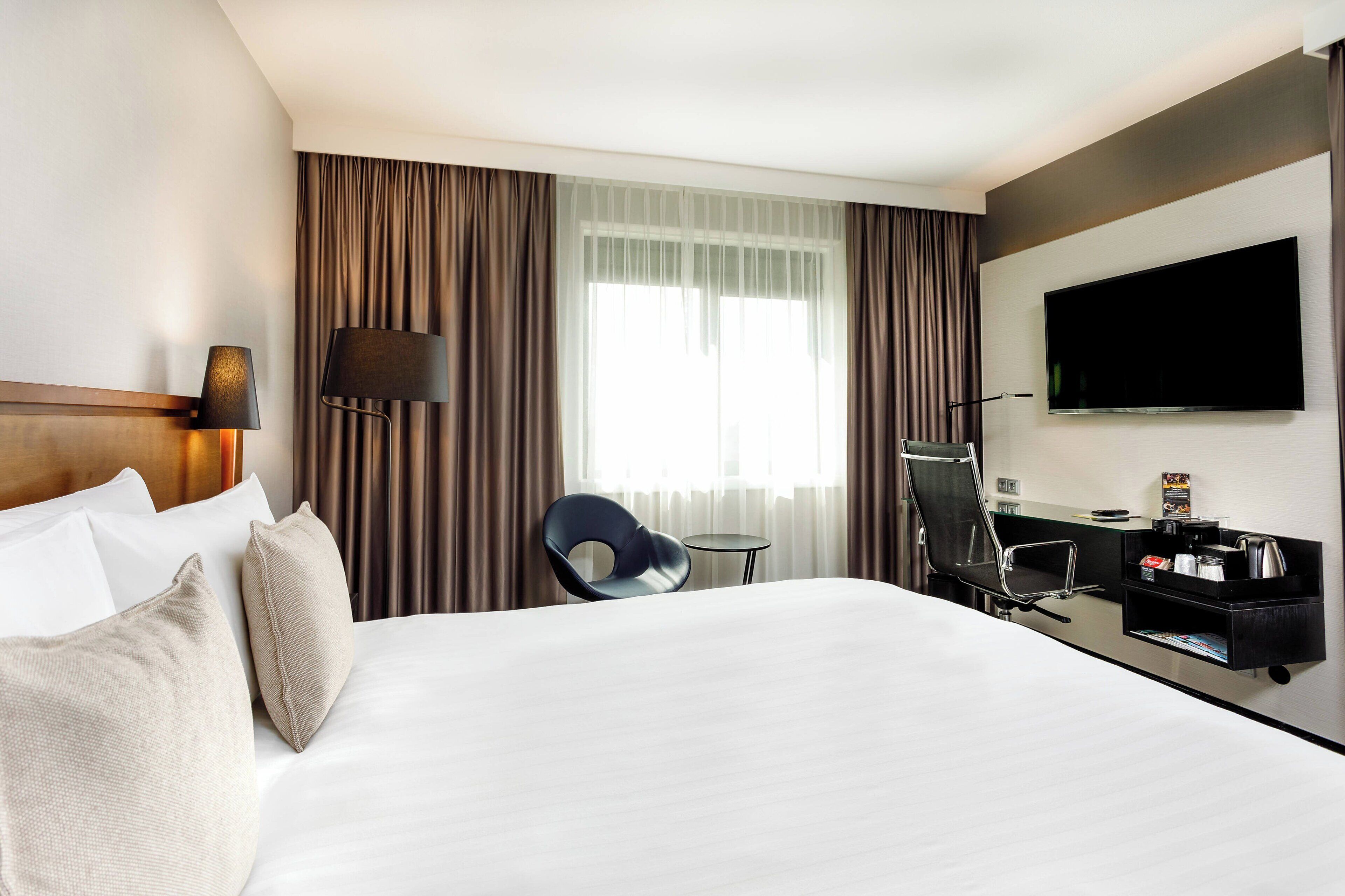 A hotel room with a large bed, a desk with a chair, a television, and a window with curtains.