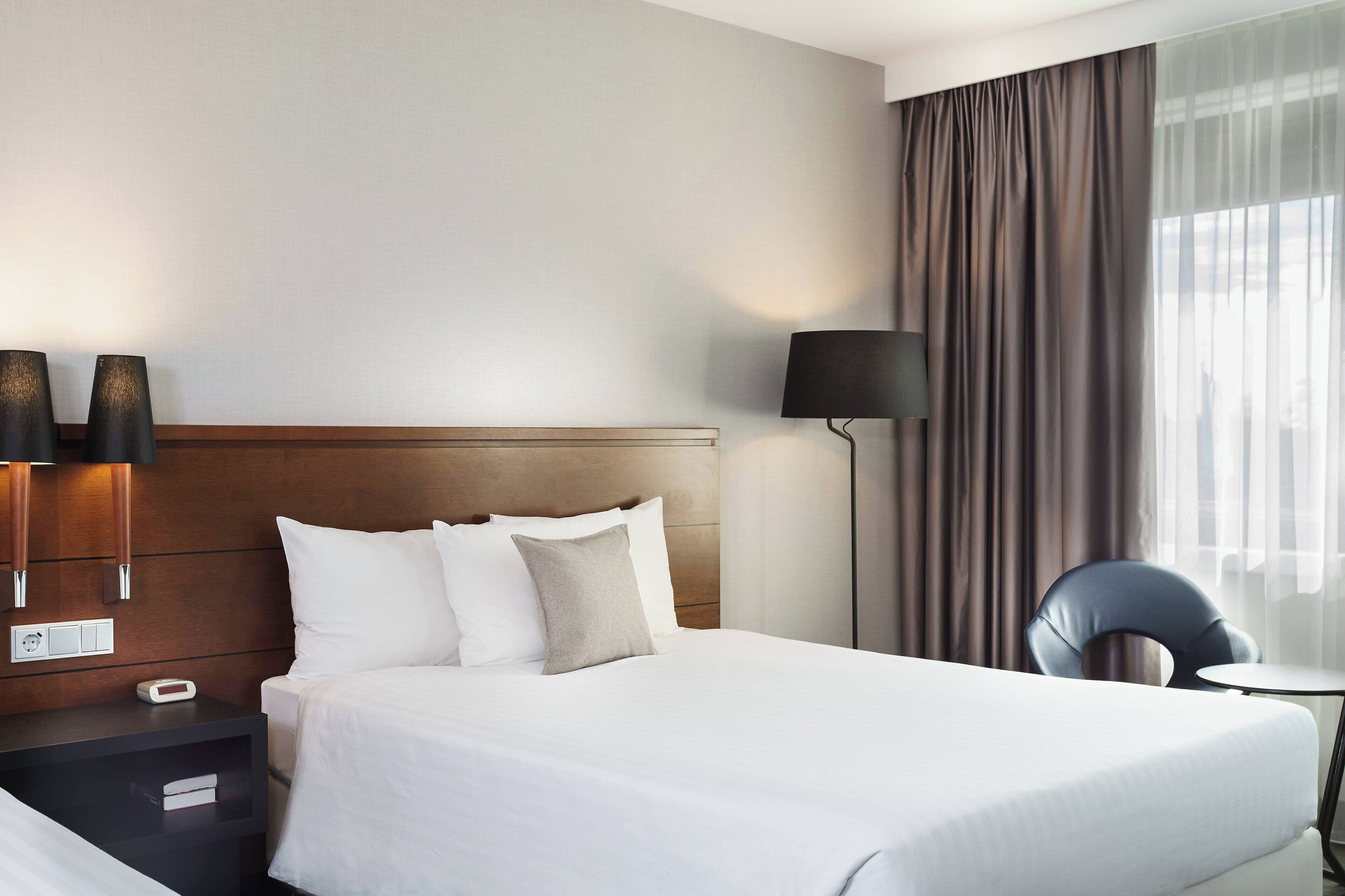 A hotel room with a bed, bedside lamps, a nightstand, a chair, and a small table.