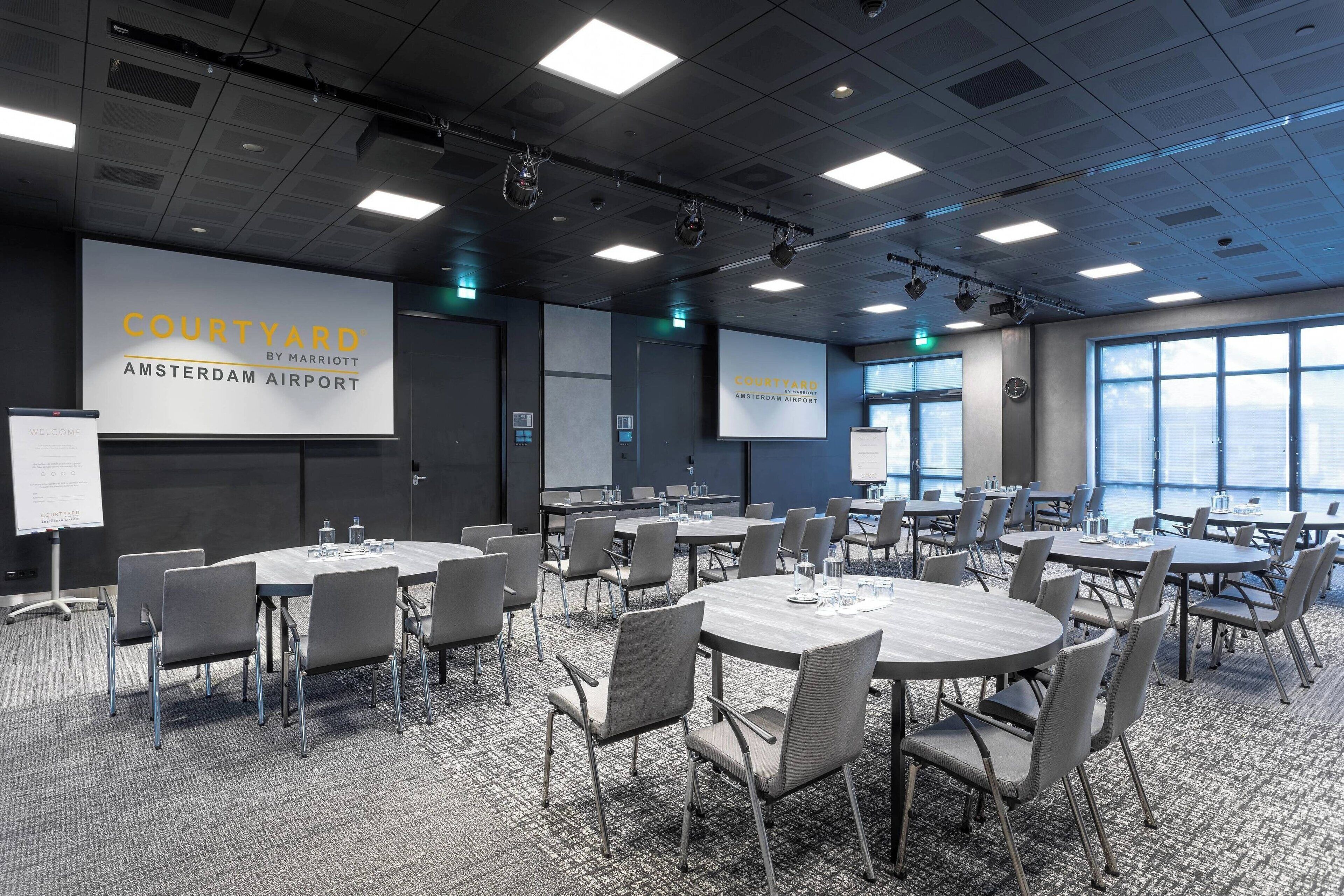 A conference room with multiple tables and chairs, large windows, and a presentation screen displaying "COURTYARD BY MARRIOTT AMSTERDAM AIRPORT".