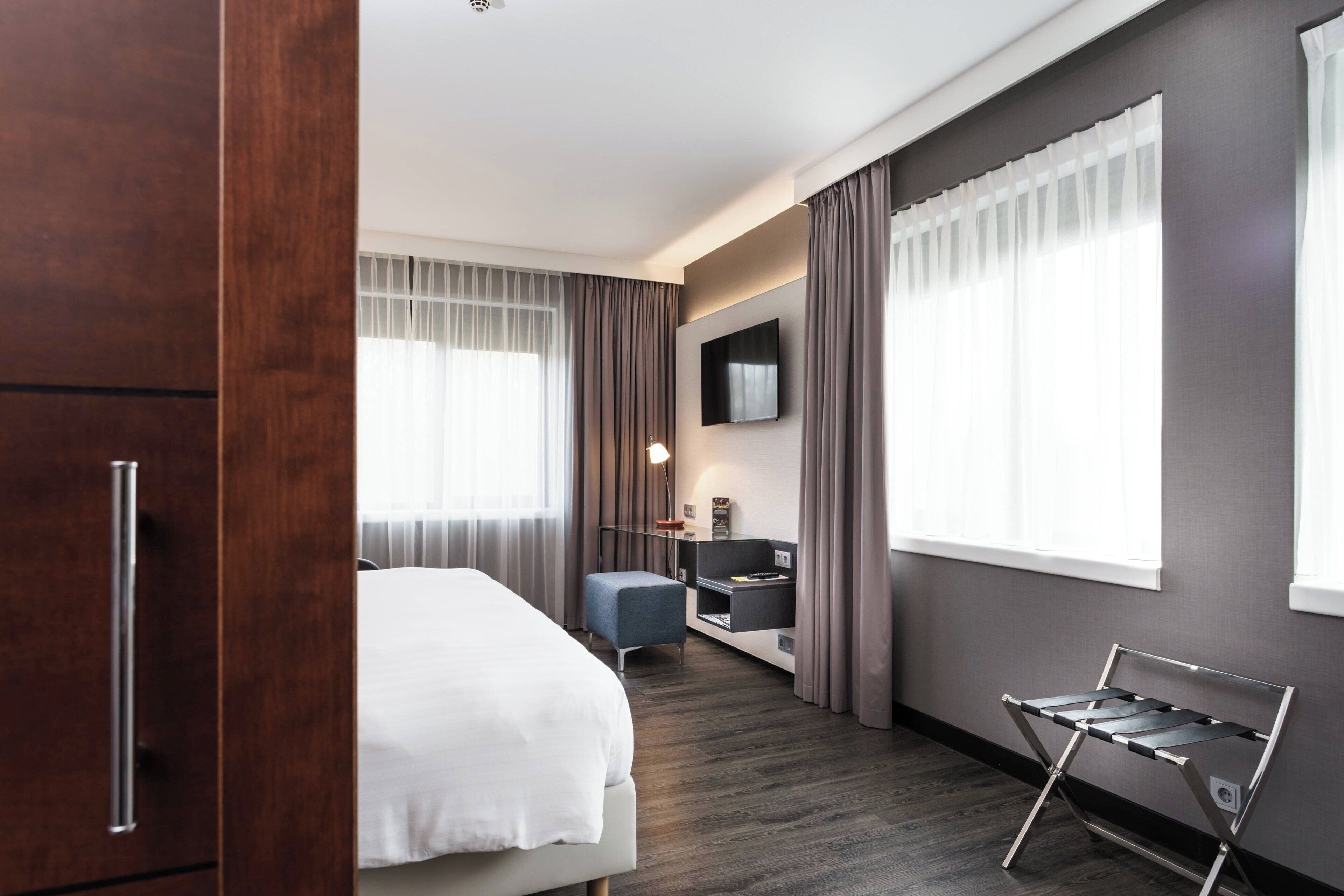 Quarto de hotel moderno com cama, escrivaninha, cadeira, televisão e janela com cortinas.
