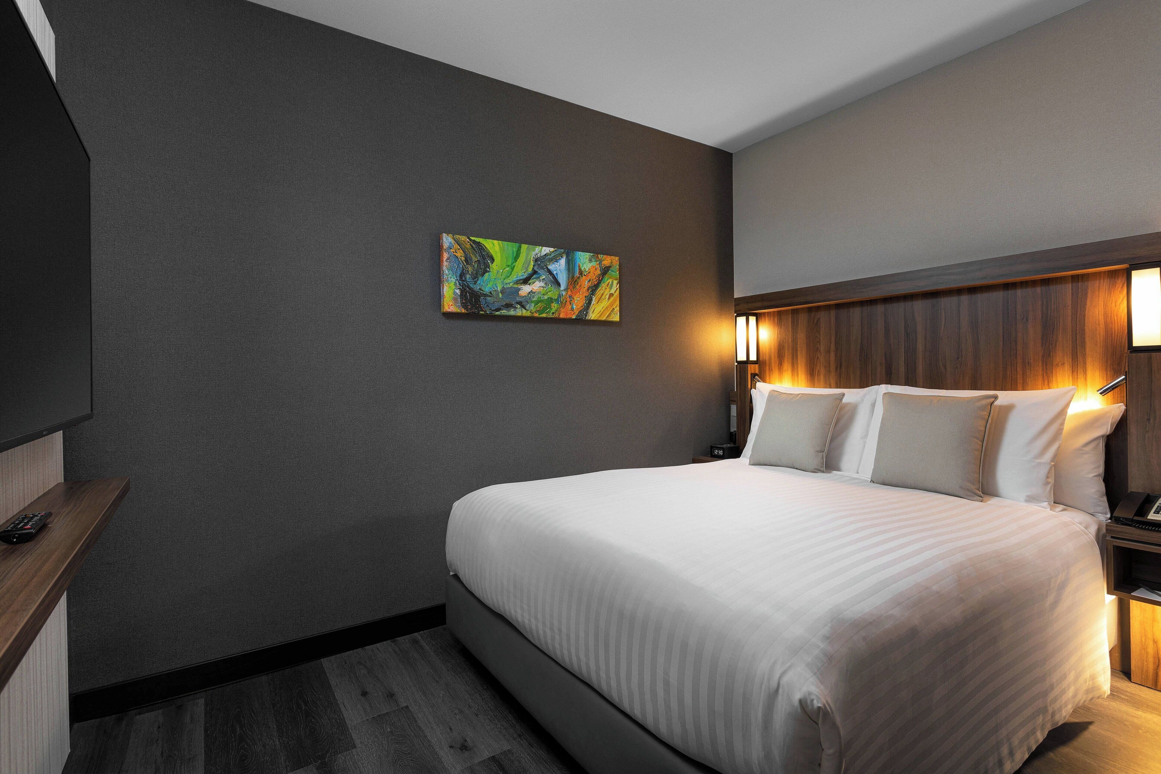 A hotel room with a large bed, a nightstand with a lamp, a painting on the wall, and a wooden headboard.