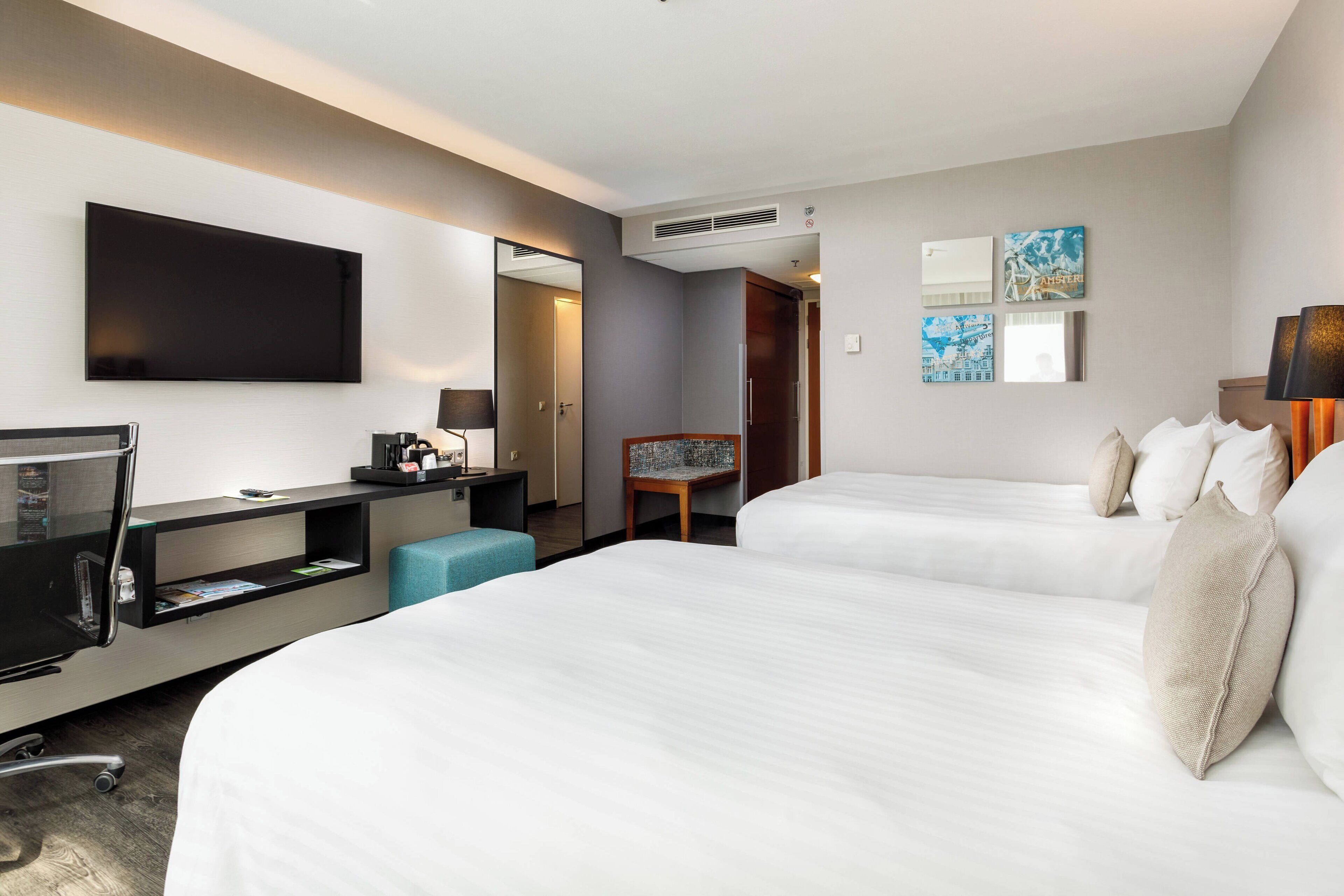 A hotel room with two beds, a desk with a flat-screen TV, a chair, and a small table.