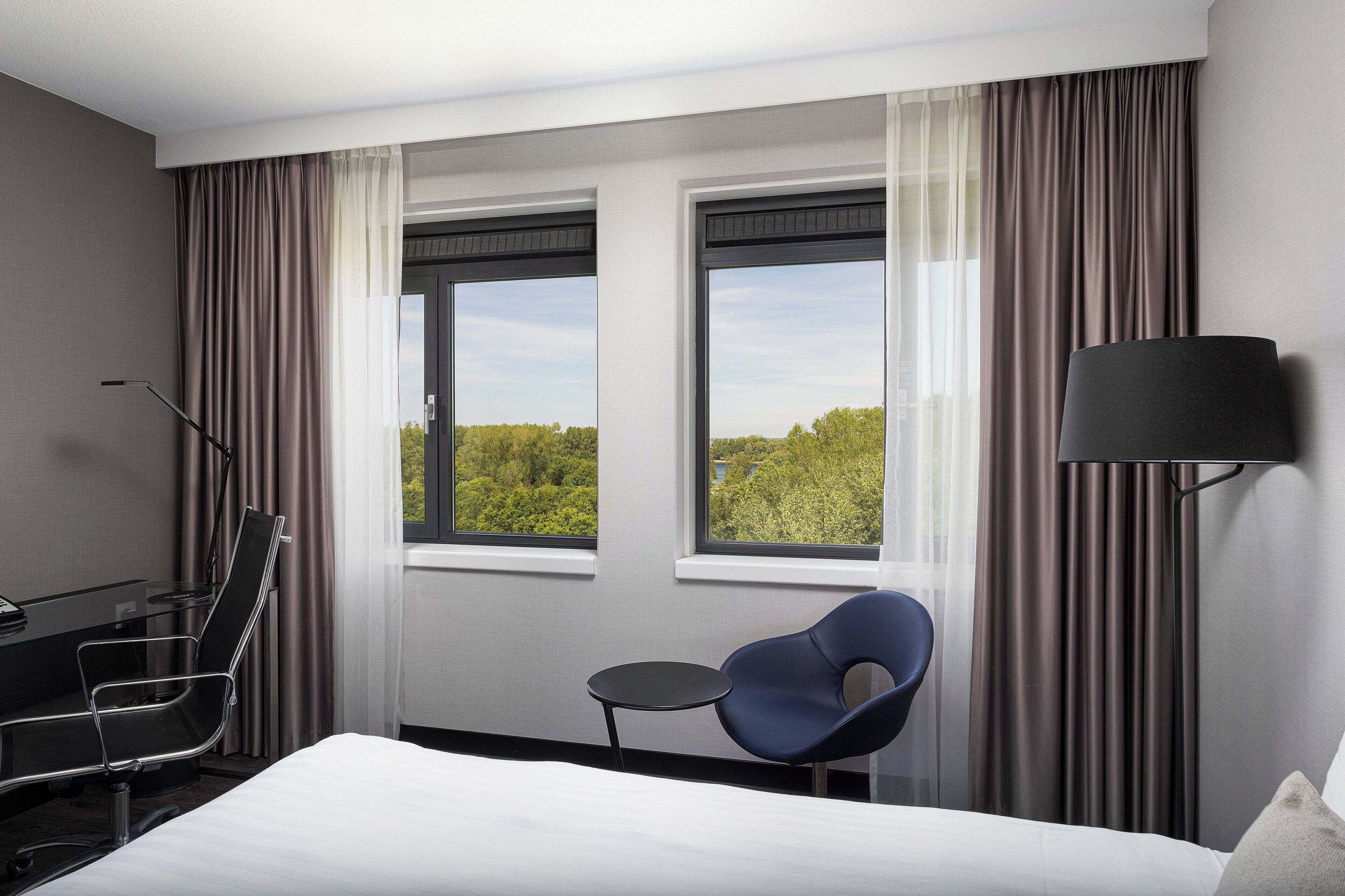 A hotel room with a bed, a desk, a chair, a small table, and two windows with curtains.