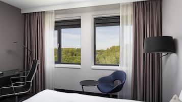 A hotel room with a bed, a desk, a chair, a small table, and two windows with curtains.