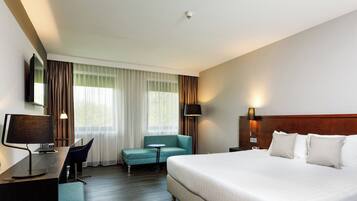 A hotel room with a bed, a desk with a lamp, a chair, a small table, and a TV on the wall.