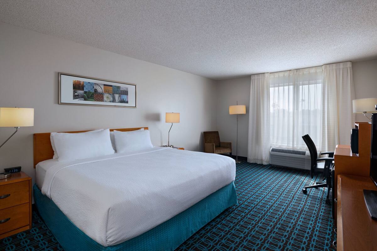 Une chambre d’hôtel avec un grand lit, deux tables de chevet, un bureau avec une chaise, une télévision et une fenêtre avec des rideaux.