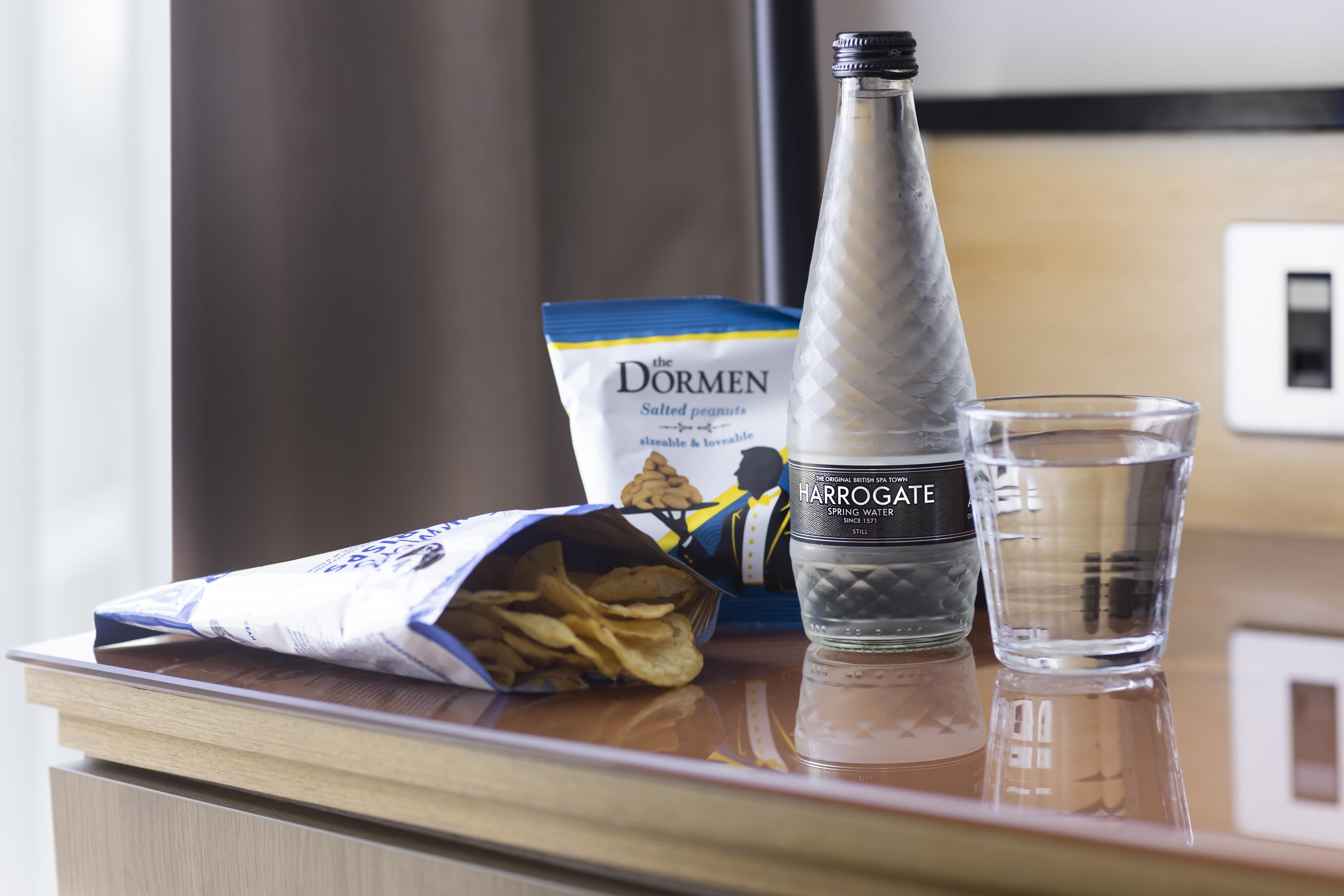 A table with a bottle of Harrogate spring water, a packet of salted peanuts, and a glass of water.