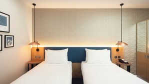 A hotel room with two single beds, a blue headboard, bedside tables with lamps, a phone, and framed pictures on the wall.