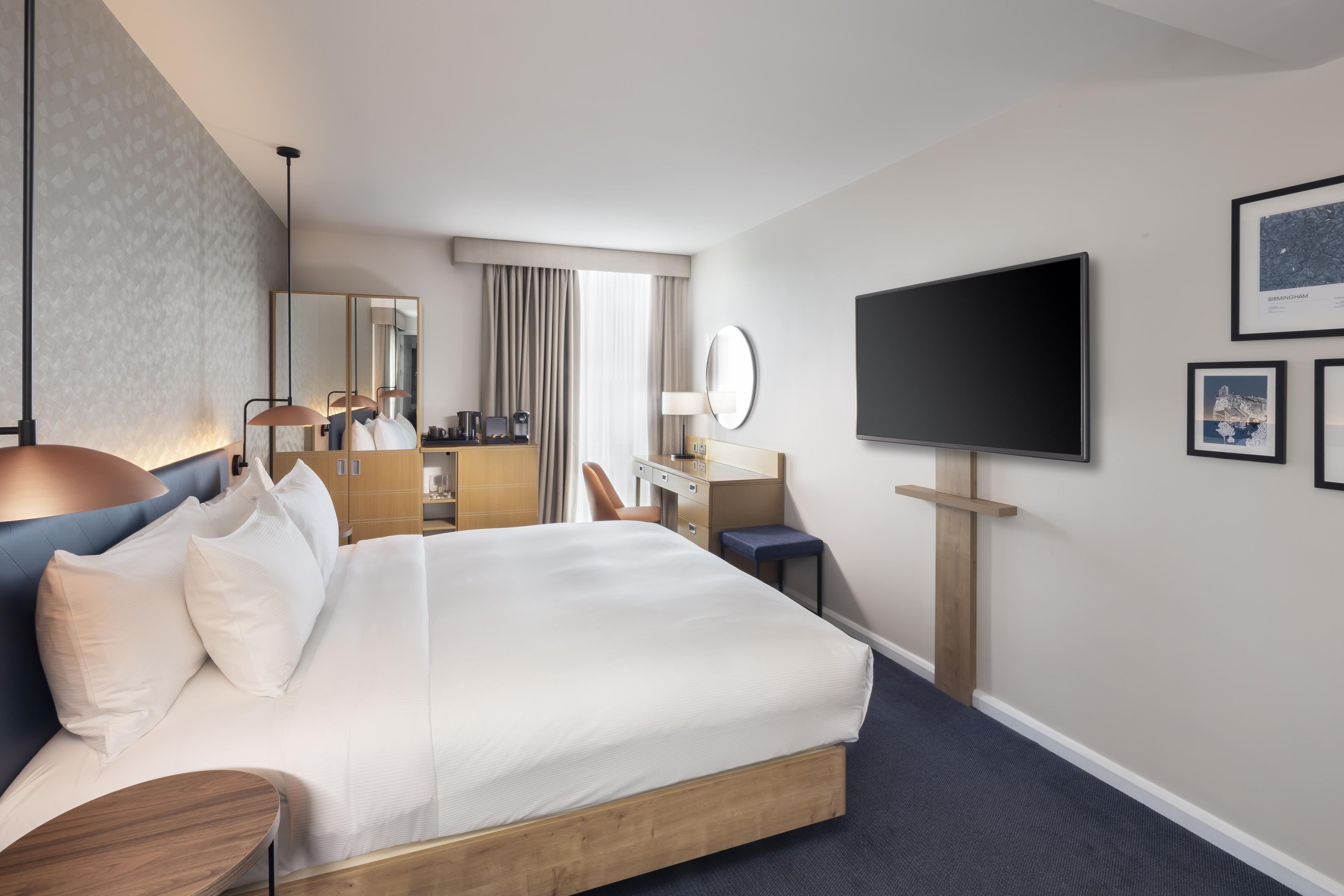A hotel room with a large bed, a desk, a chair, a flat-screen TV, and a mirror.