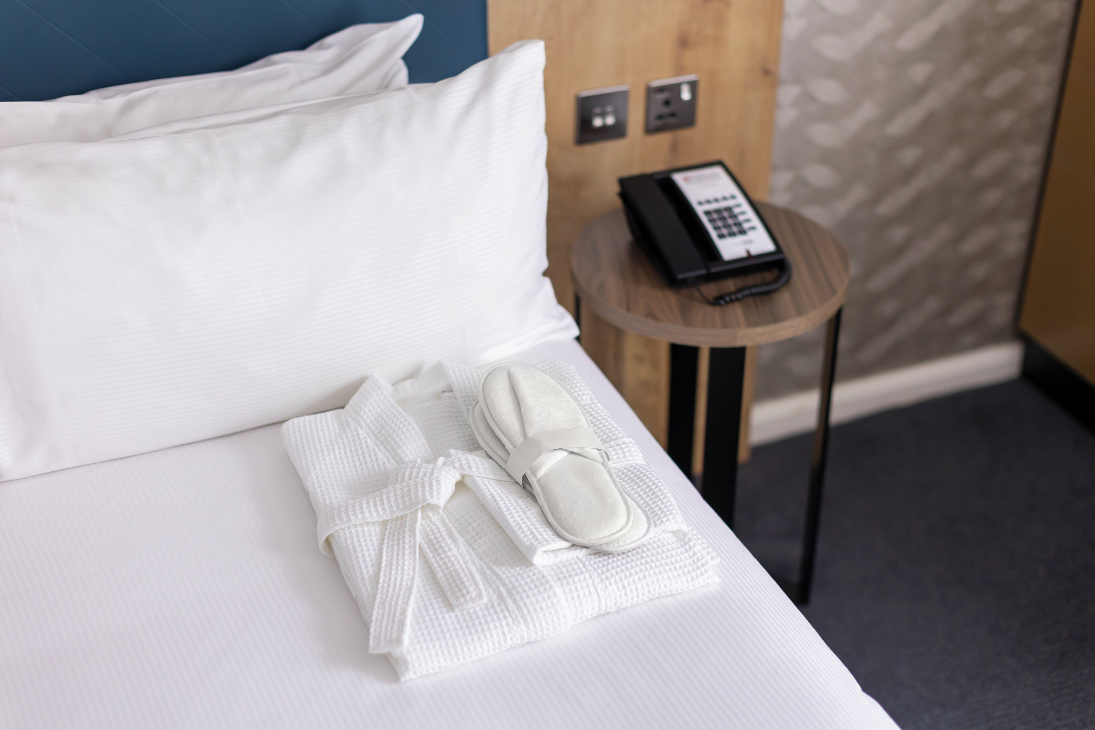 A neatly made bed with white linens, a pair of slippers, and a folded towel on a wooden bedside table with a telephone.