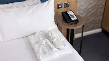 A neatly made bed with white linens, a pair of slippers, and a folded towel on a wooden bedside table with a telephone.