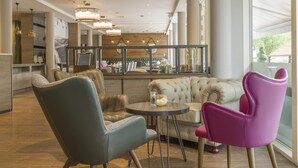 A modern interior with a seating area featuring a tufted sofa and two chairs, a round table with a glass, and a large mirror reflecting the room. - Hilton Garden Inn Bristol City Centre (Bristol)