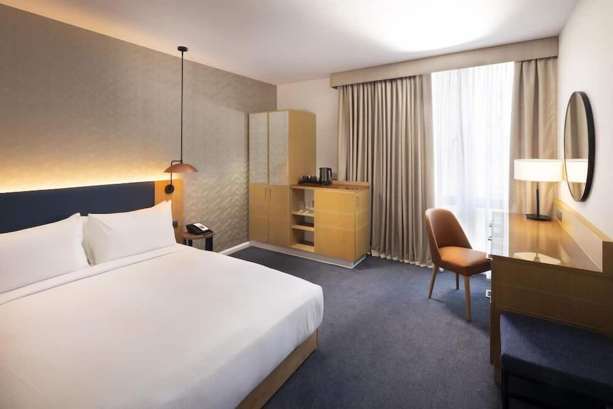 Une chambre d’hôtel comprenant un lit, un bureau, une chaise, une commode, un miroir et une fenêtre avec des rideaux.