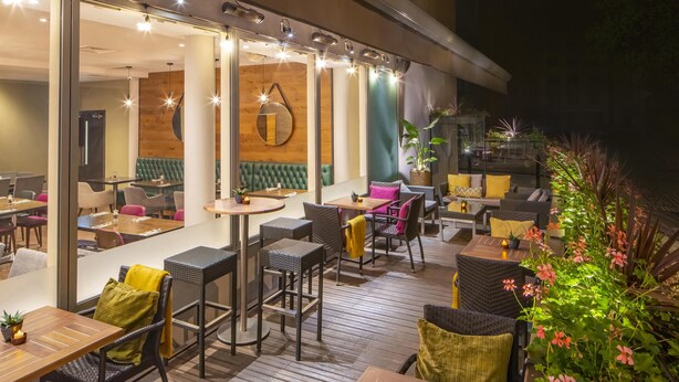 An outdoor dining area with tables and chairs, a wooden deck, and potted plants. - Hilton Garden Inn Bristol City Centre (Bristol)