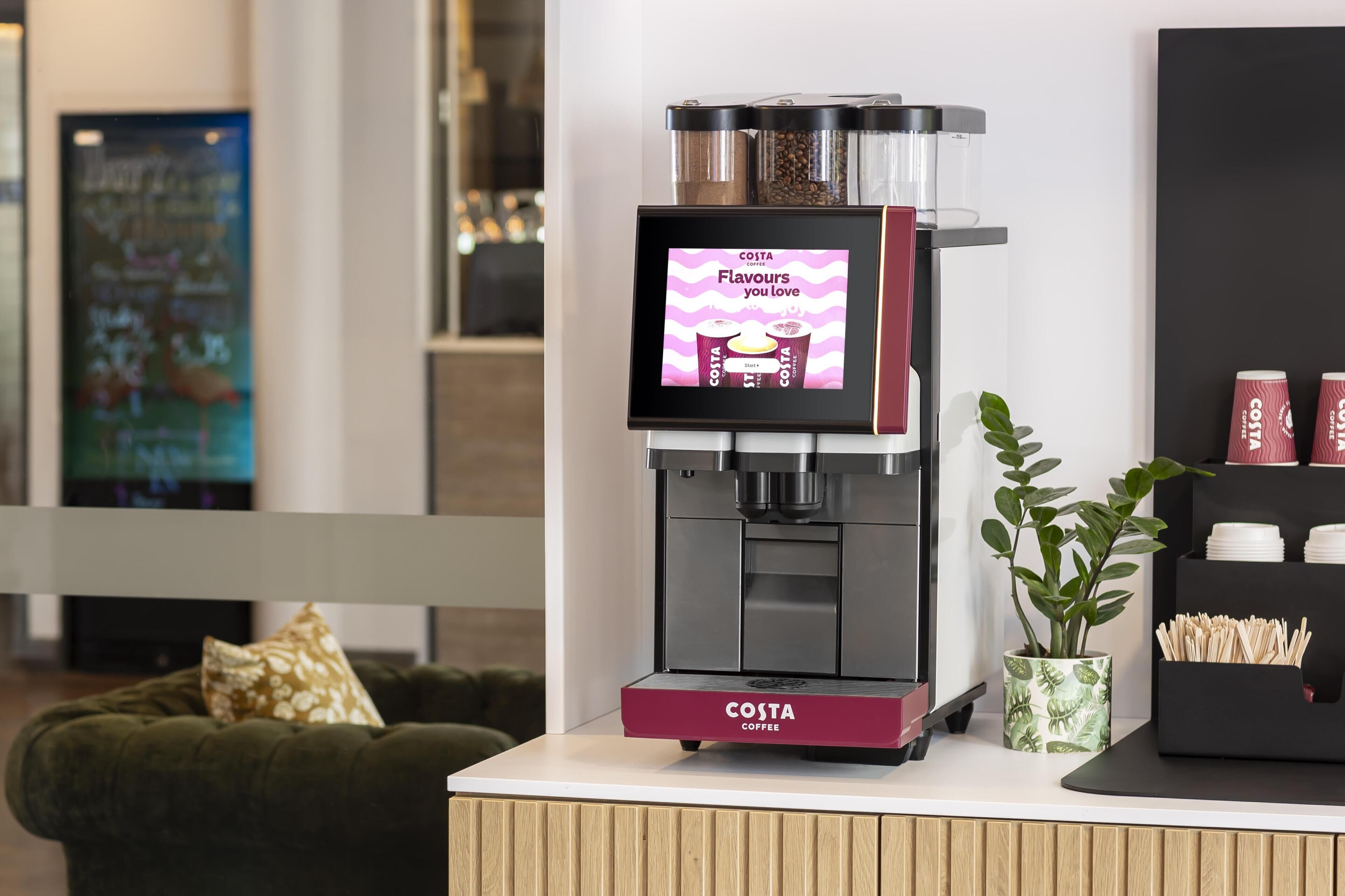 A Costa Coffee branded coffee machine with a digital display, a plant, and a stack of disposable cups.