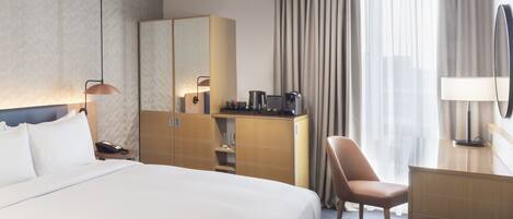 Habitación de hotel con una cama grande, un escritorio con silla, armario, espejo y ventana con cortinas.