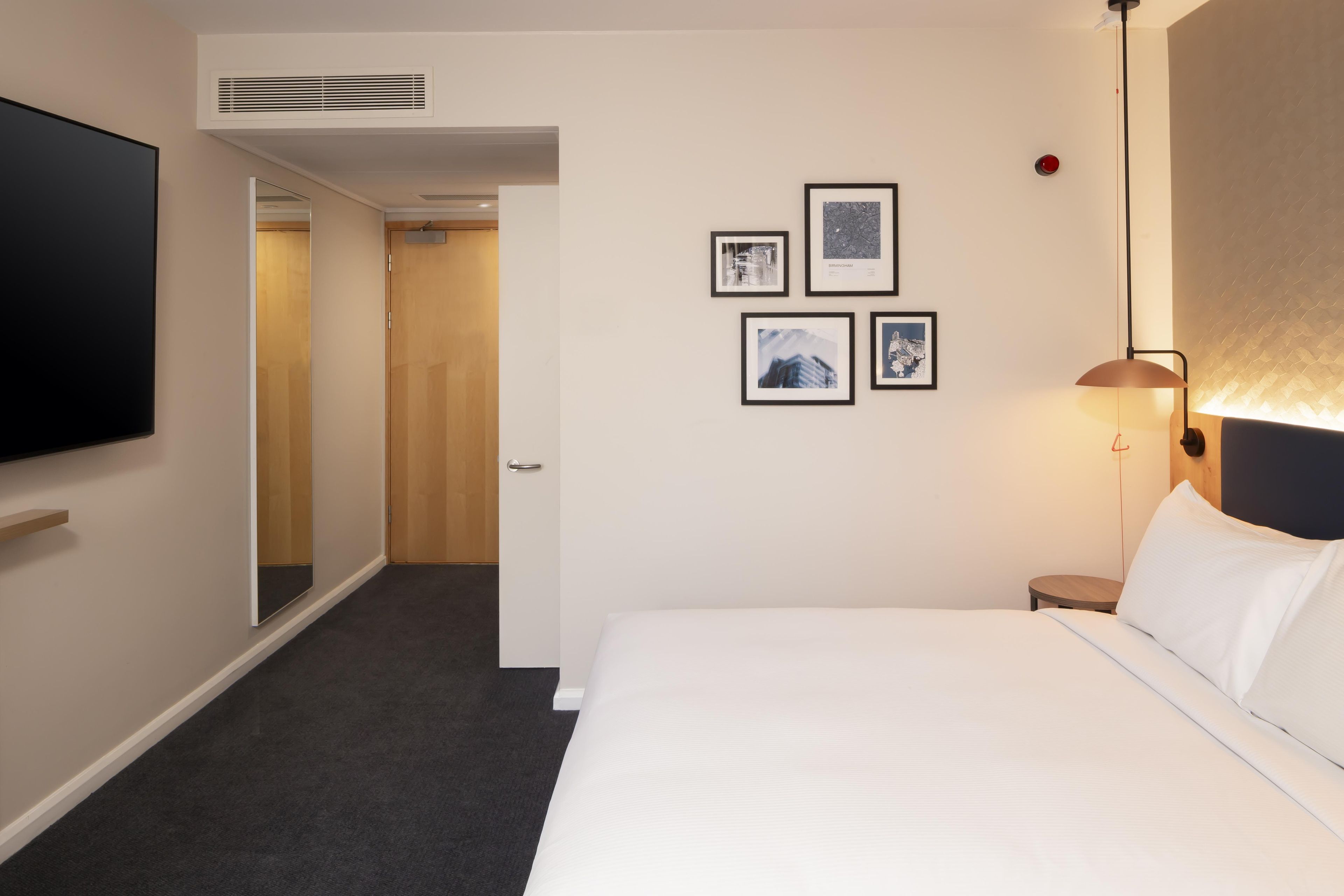 A hotel room with a bed, a TV, a wall-mounted lamp, and framed pictures.