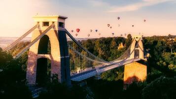 A suspension bridge with a tower structure, surrounded by greenery and a clear sky.